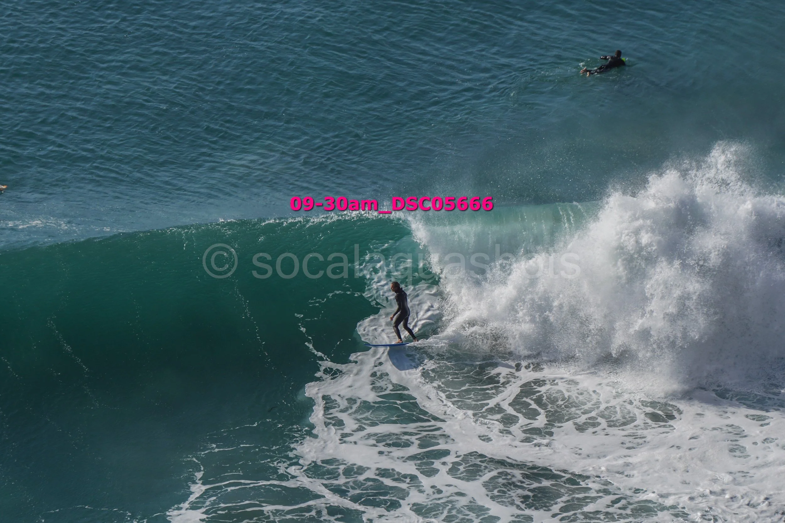 A person surfing on a large ocean wave with a surfer in a wetsuit in the background.