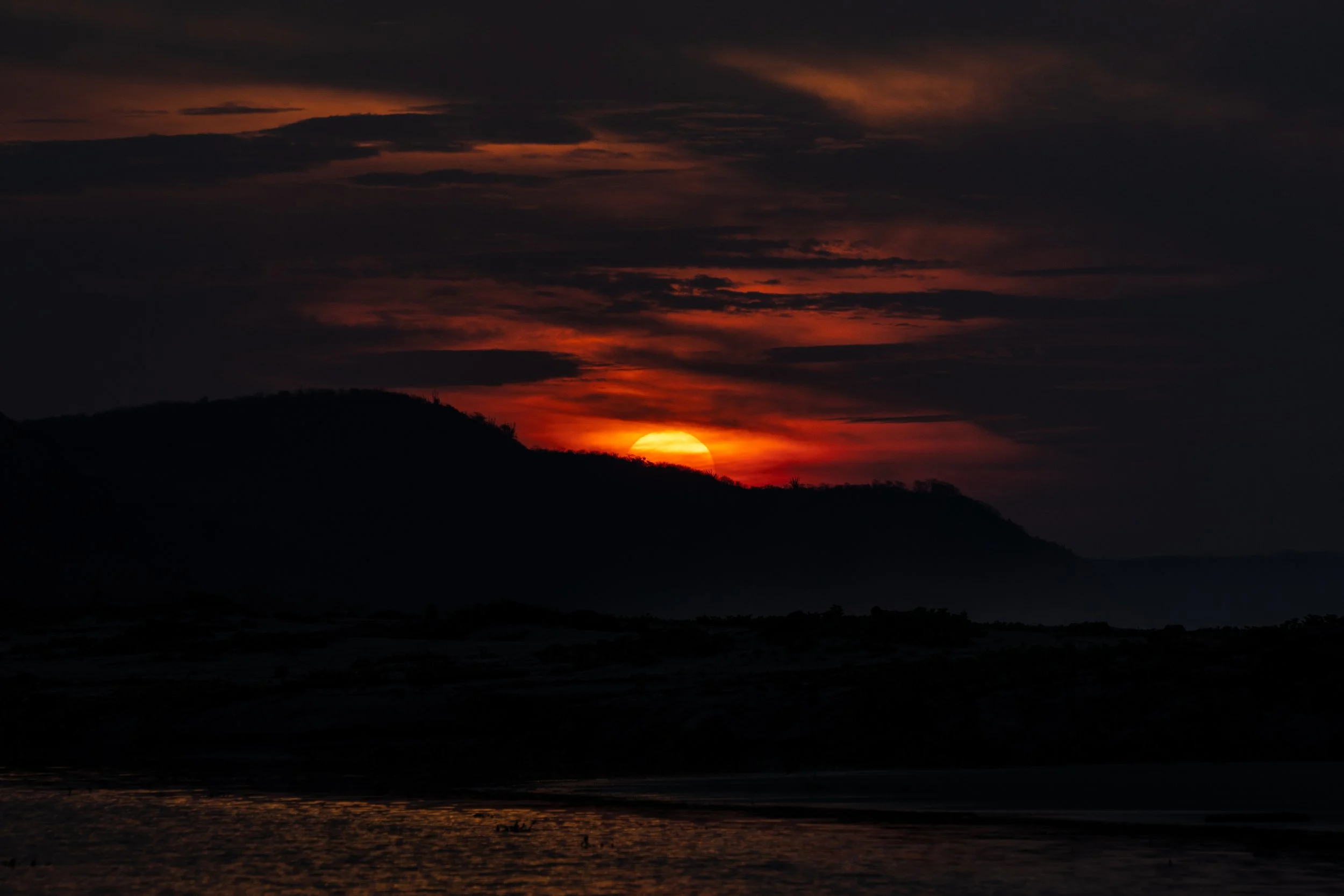 Sunset over a mountainous landscape with dark clouds and a partially visible setting sun, with reflections on water.