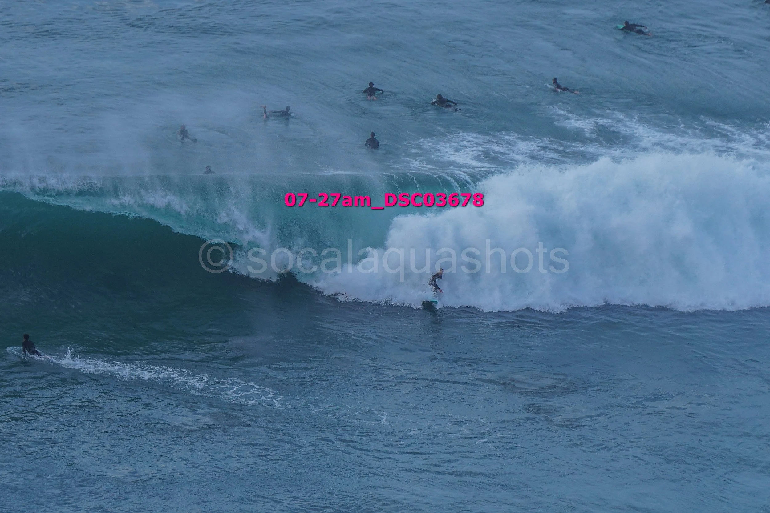 Surfers riding large ocean waves with many people in the water in the background.
