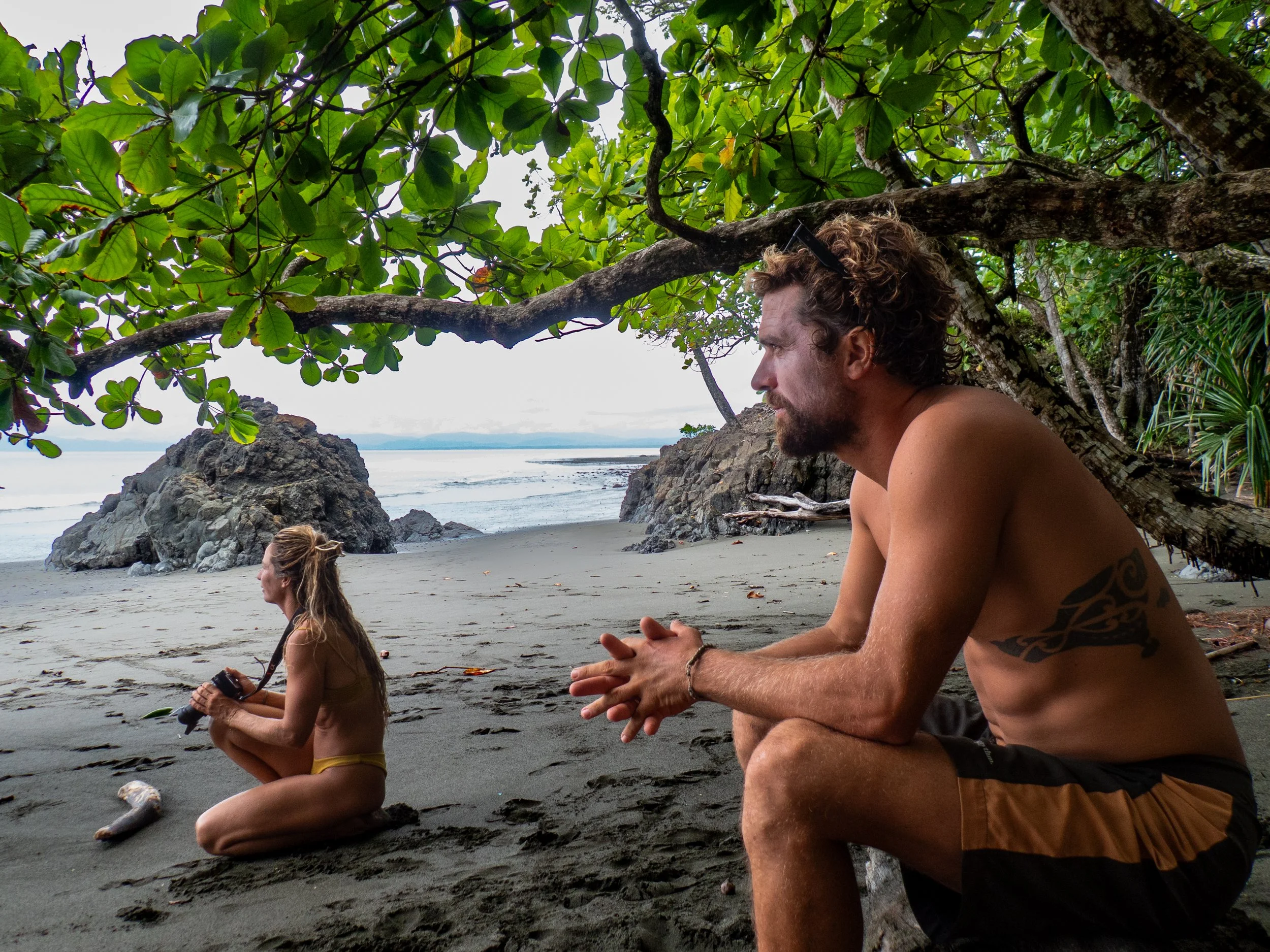 A man and woman meditating on the beach under a tree with green leaves, rocks, and the ocean in the background.