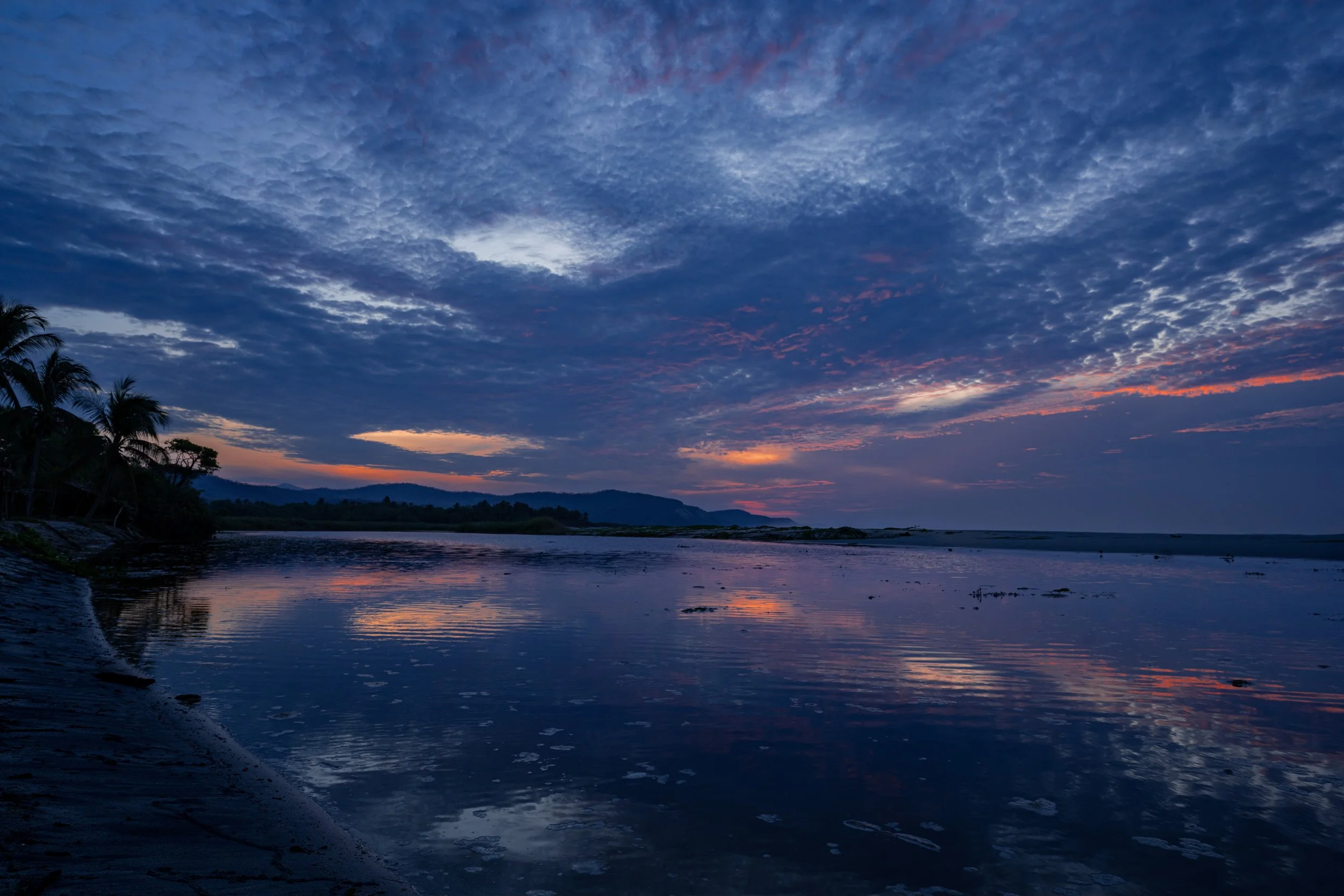 A landscape scene of a river at dusk with a partly cloudy sky, silhouettes of trees on the left, and distant mountains on the horizon.