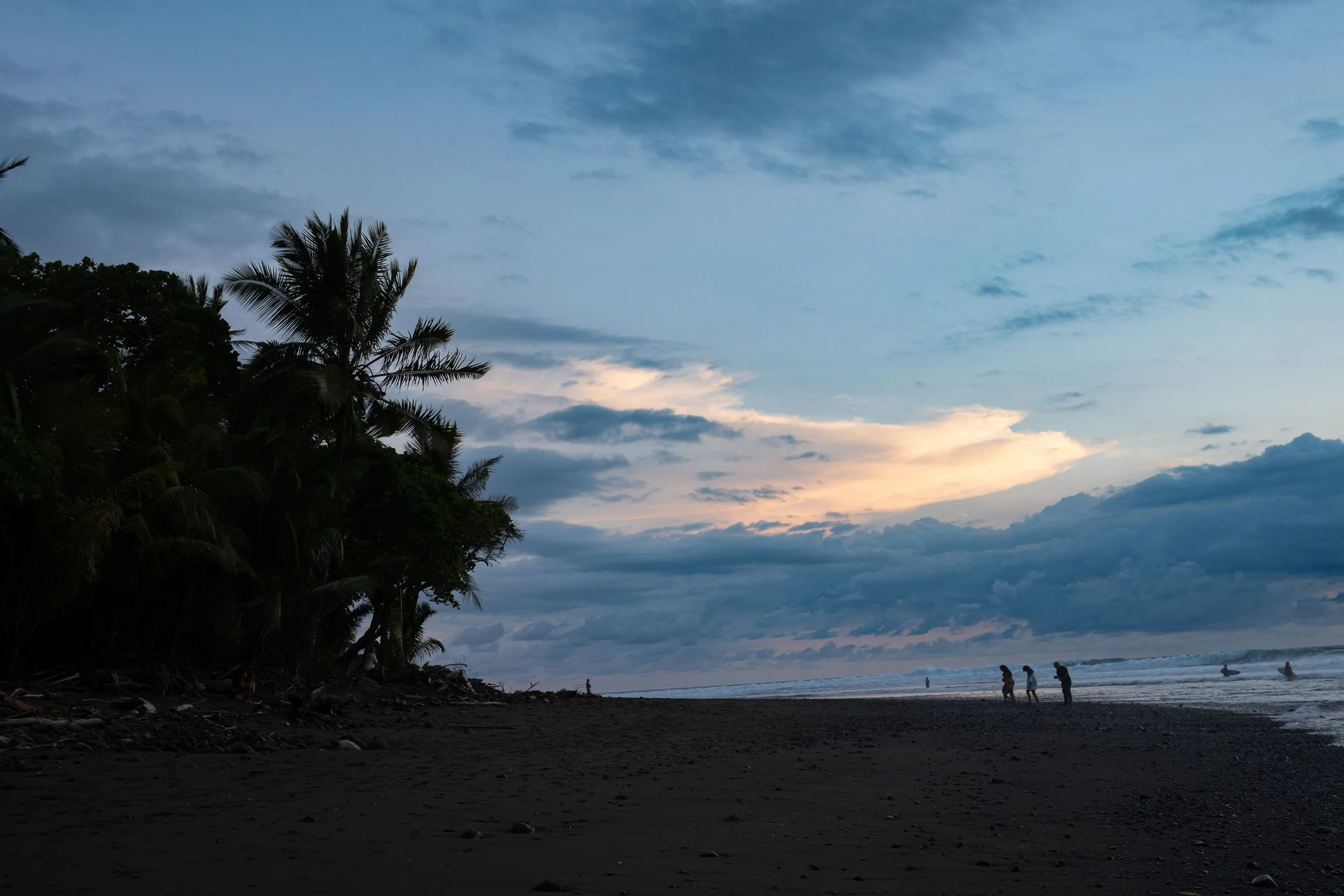 A beach at dusk with silhouettes of people near the water, lush trees on the left, and a partly cloudy sky with hints of sunset.