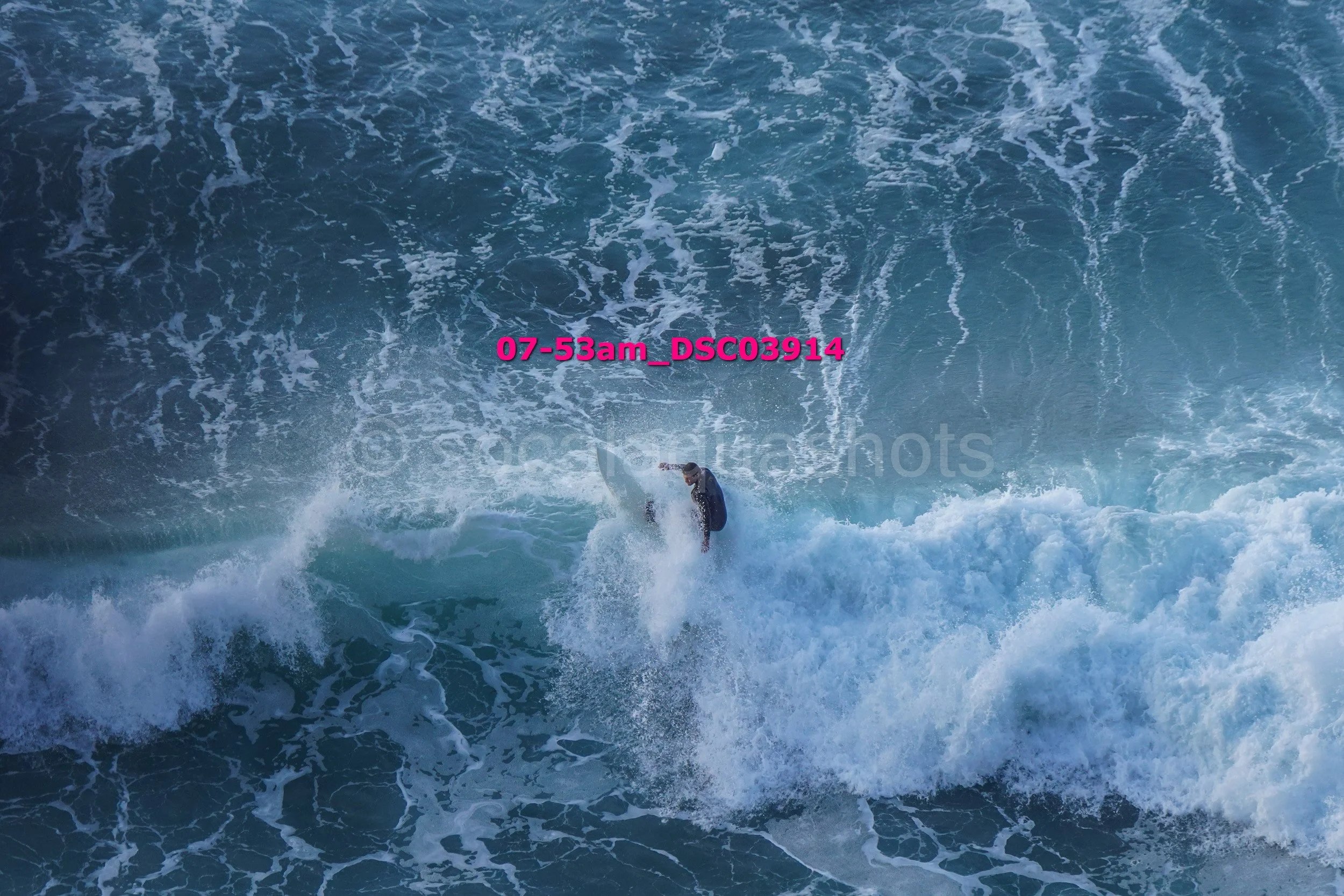 A surfer in a wetsuit riding a wave in the ocean with white foam and turquoise water, viewed from above.