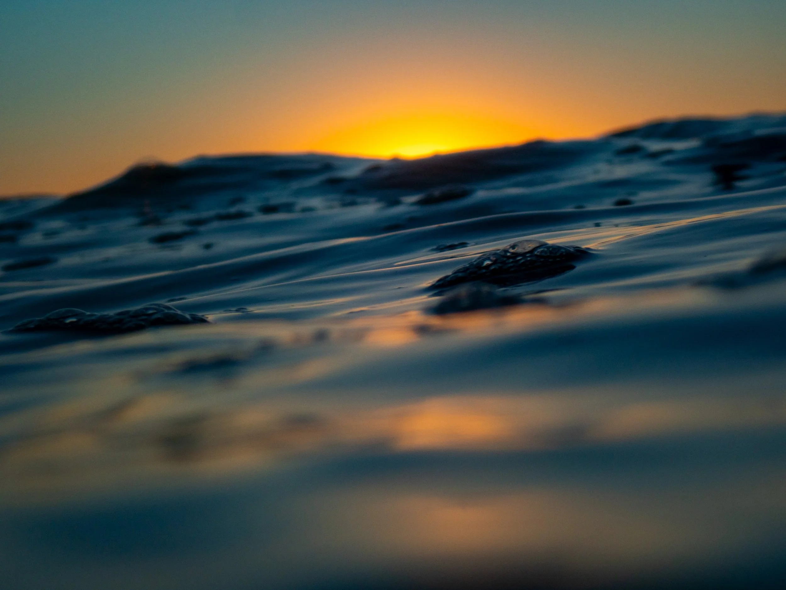 Close-up of ocean waves during sunset or sunrise, with the sun partially visible on the horizon and casting a warm glow over the water.