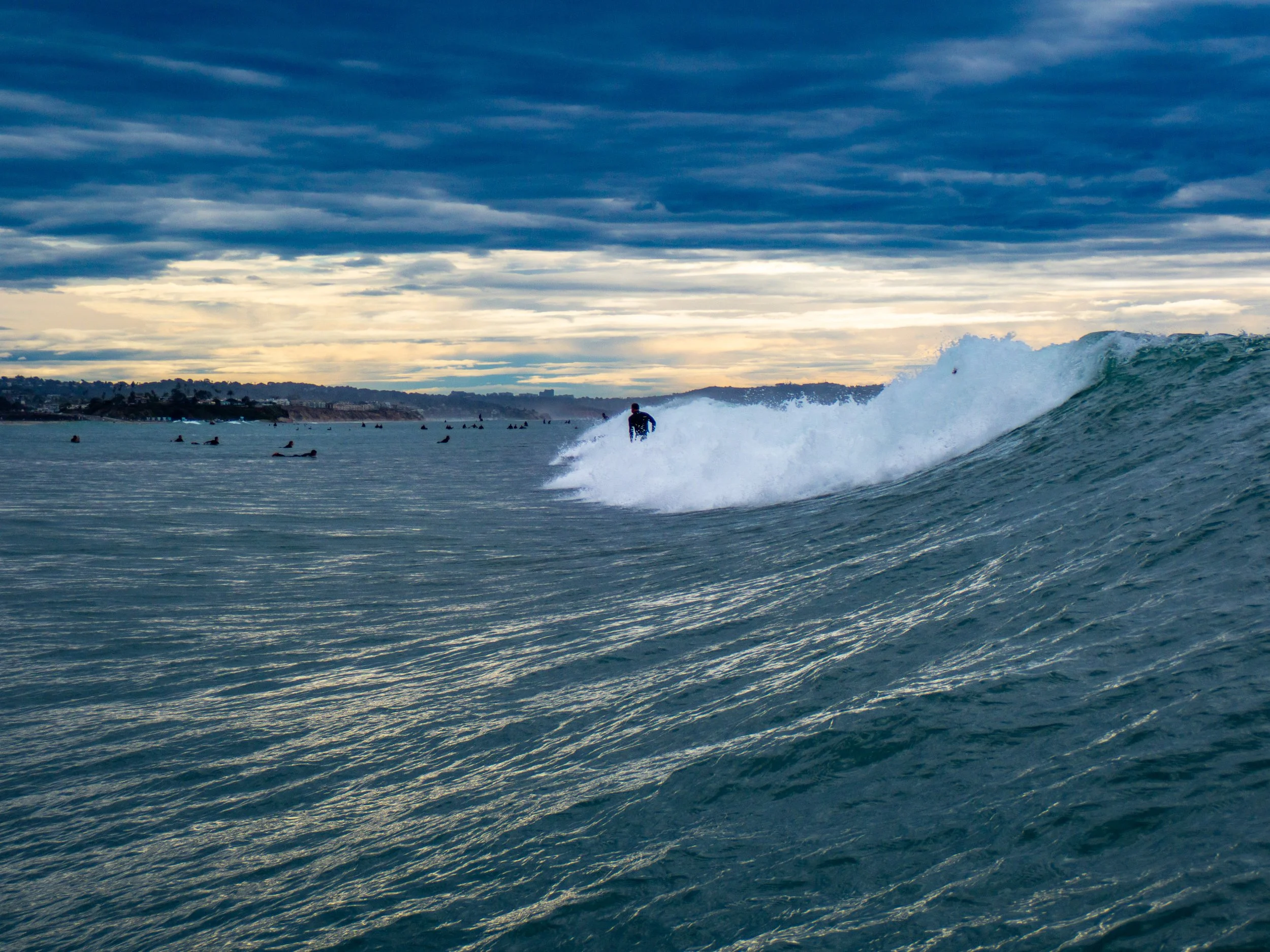 A person surfing on a wave in the ocean during cloudy weather with a coastline and multiple surfers in the background.
