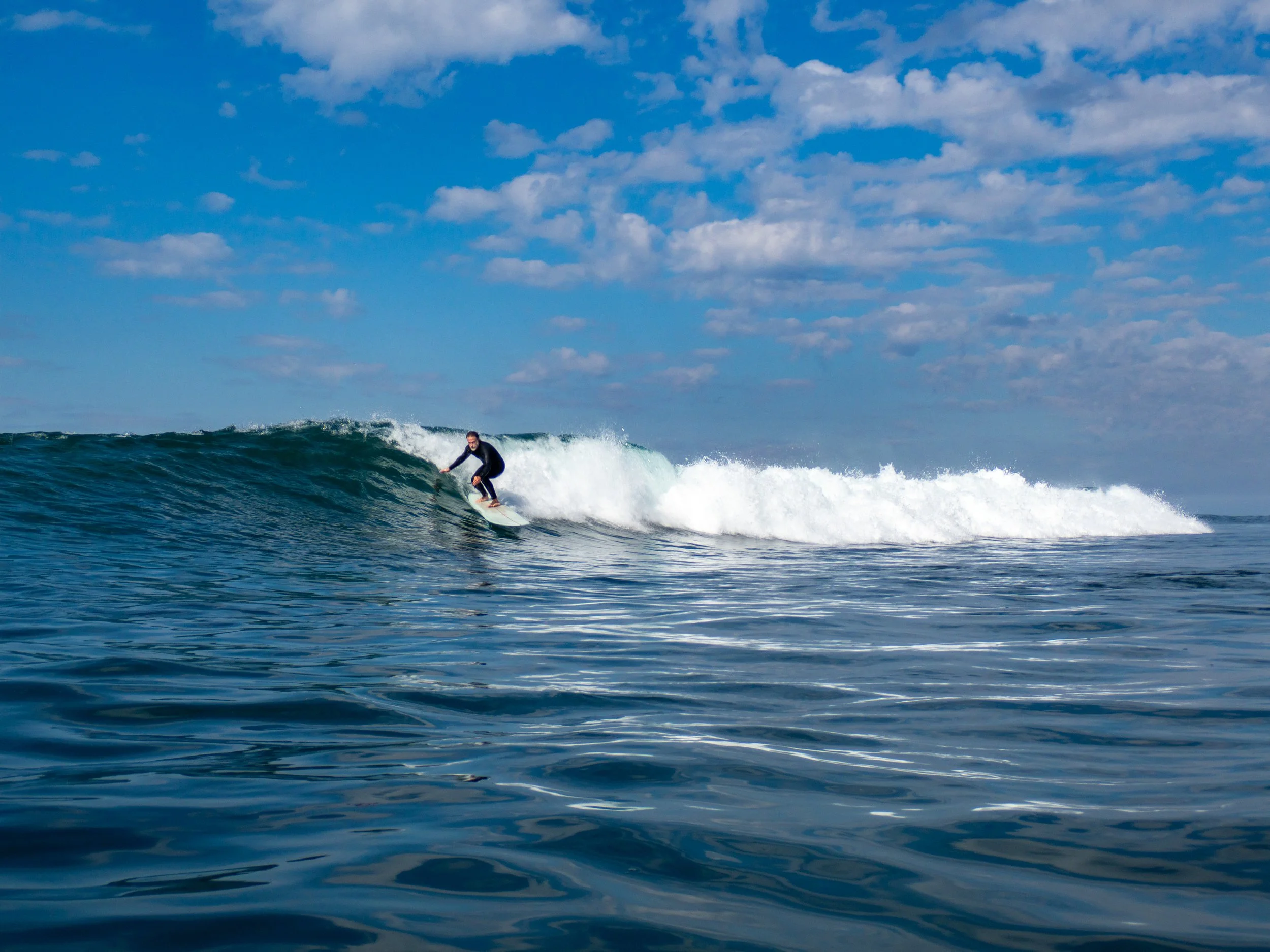A person riding a surfboard on a wave in the ocean under a blue sky with scattered clouds.