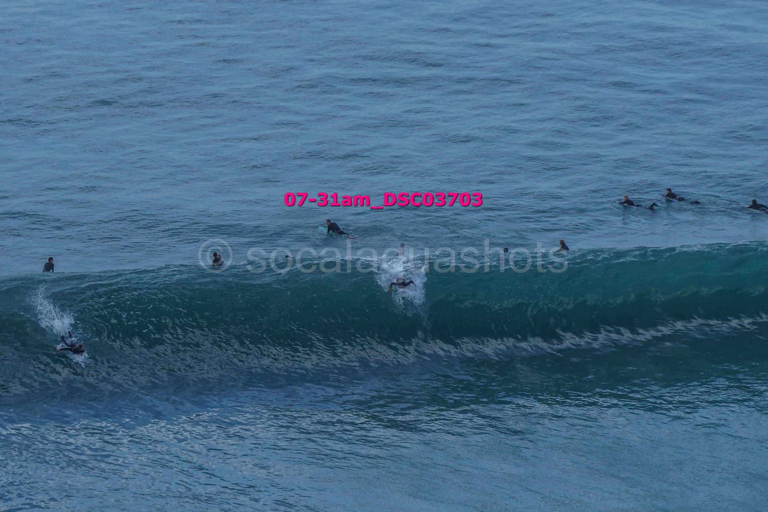 A group of surfers in wetsuits waiting in the ocean, with one surfer catching a wave and falling into the water.