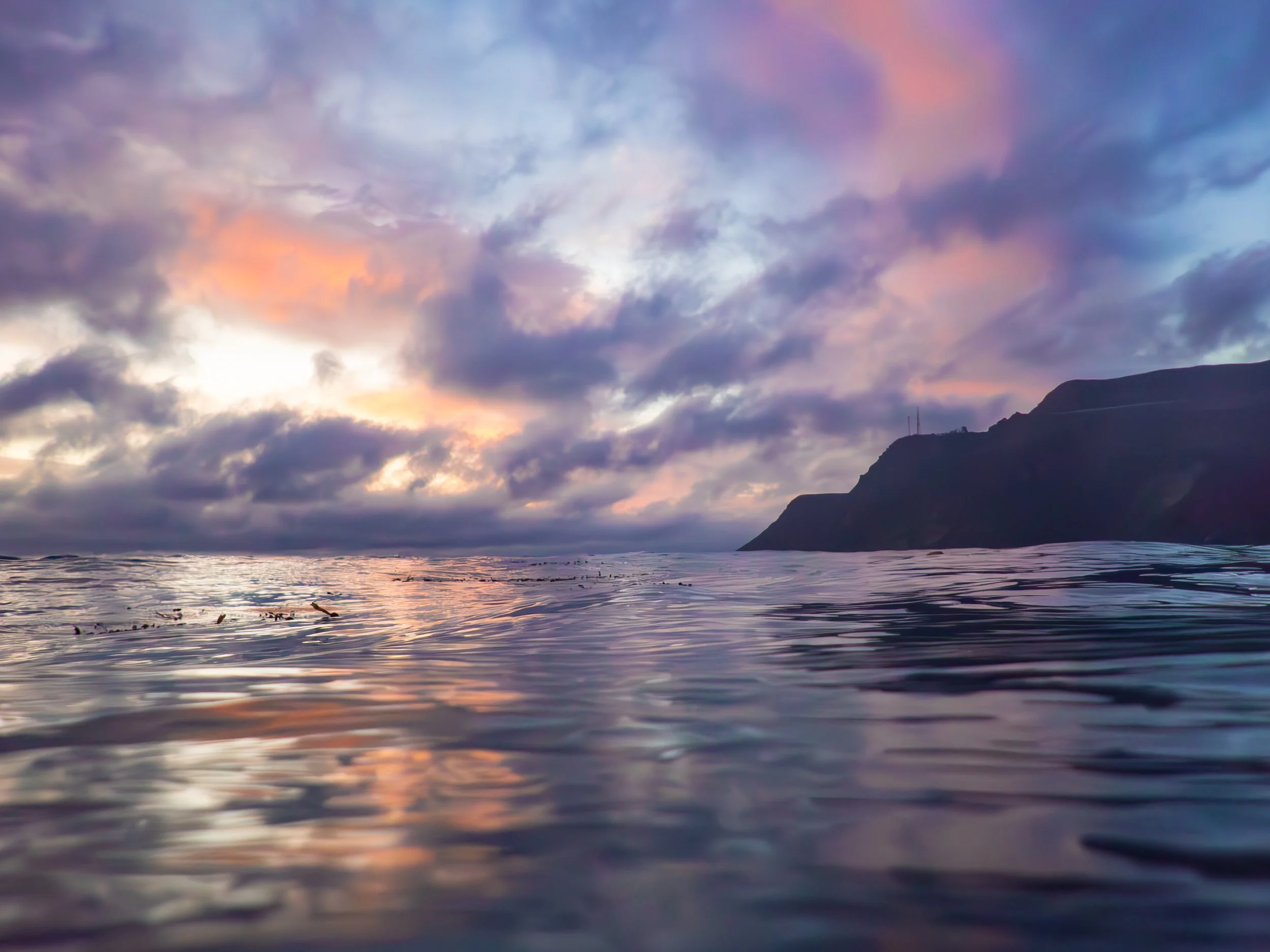 A view of the ocean at sunset with a partly cloudy sky and a silhouette of a mountain in the distance.