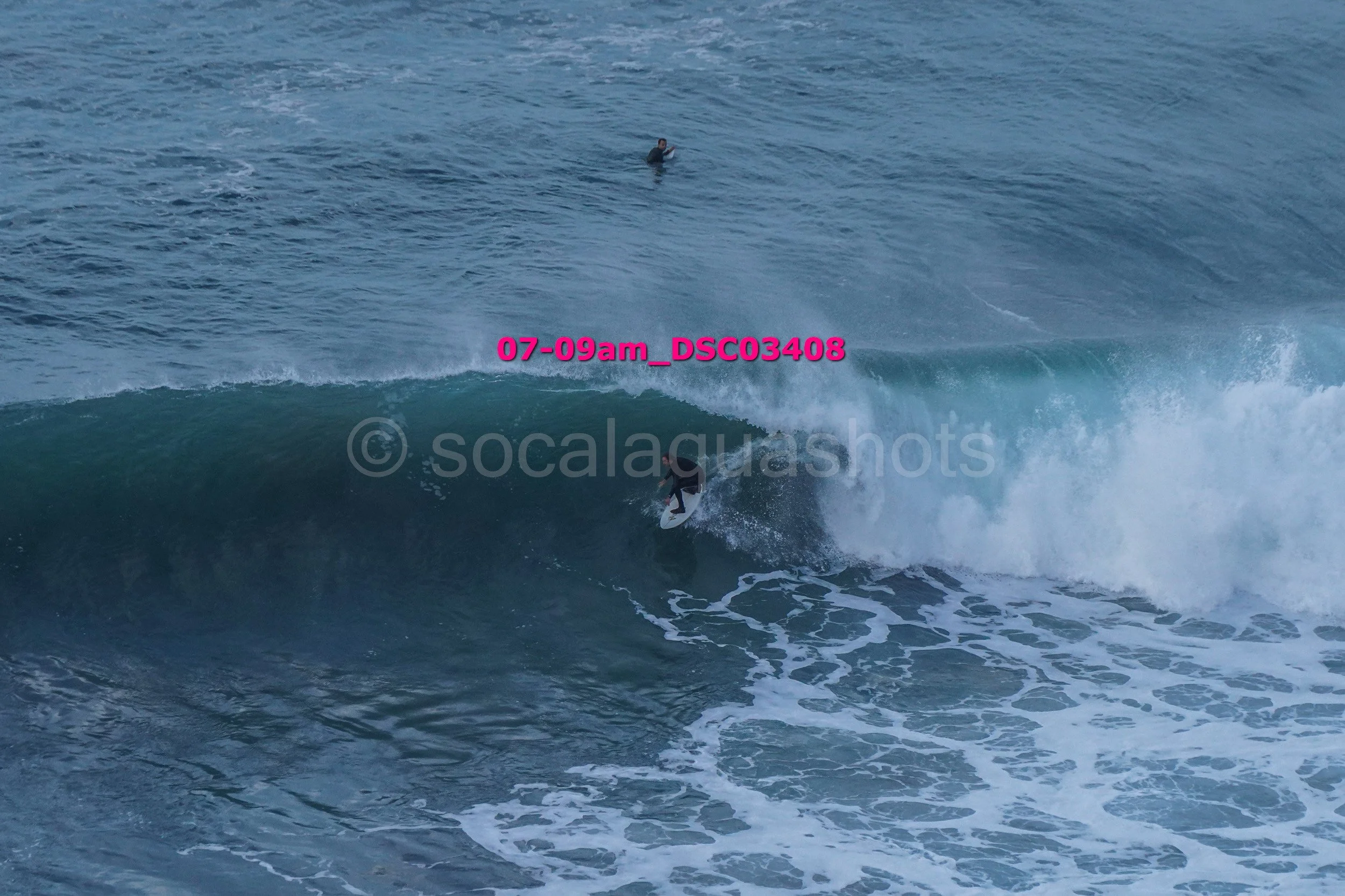 A surfer riding a large ocean wave with another person swimming in the distance.