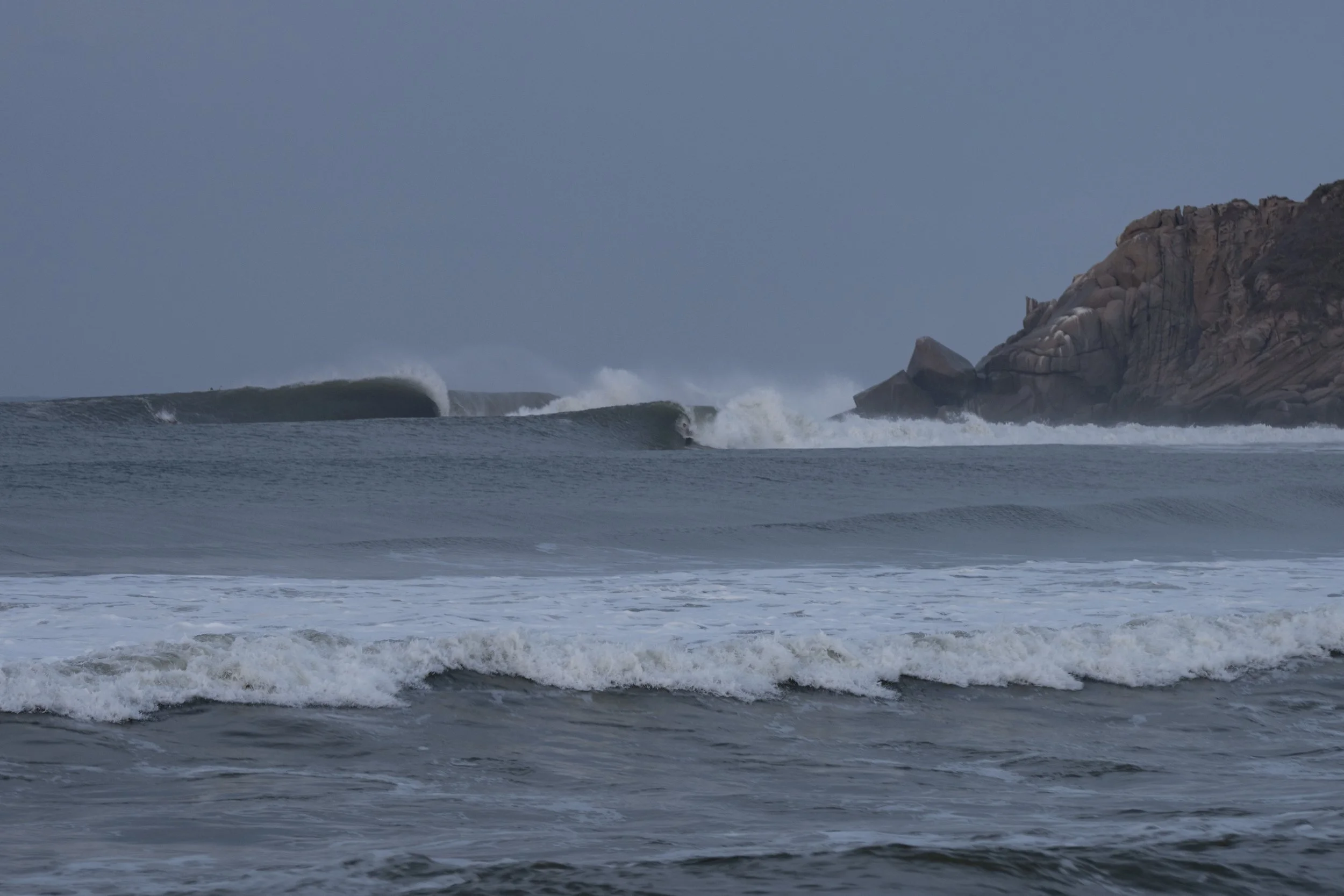 Ocean waves crashing against the shoreline near rocky cliffs under a cloudy sky.