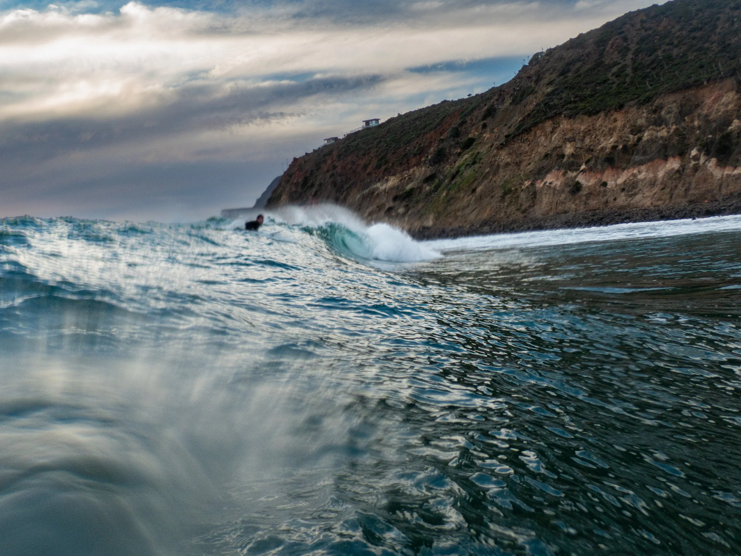 A person surfing on a wave near a rocky coastline with hills and overcast sky.