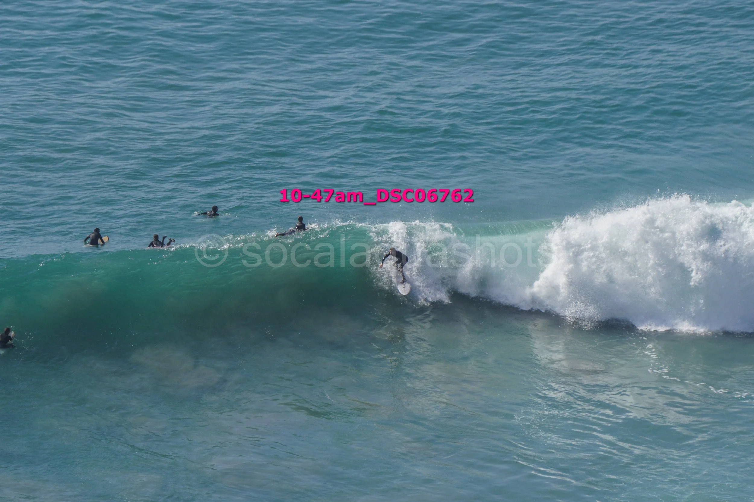 A surfer riding a wave with several people in the water watching, in a large ocean scene.