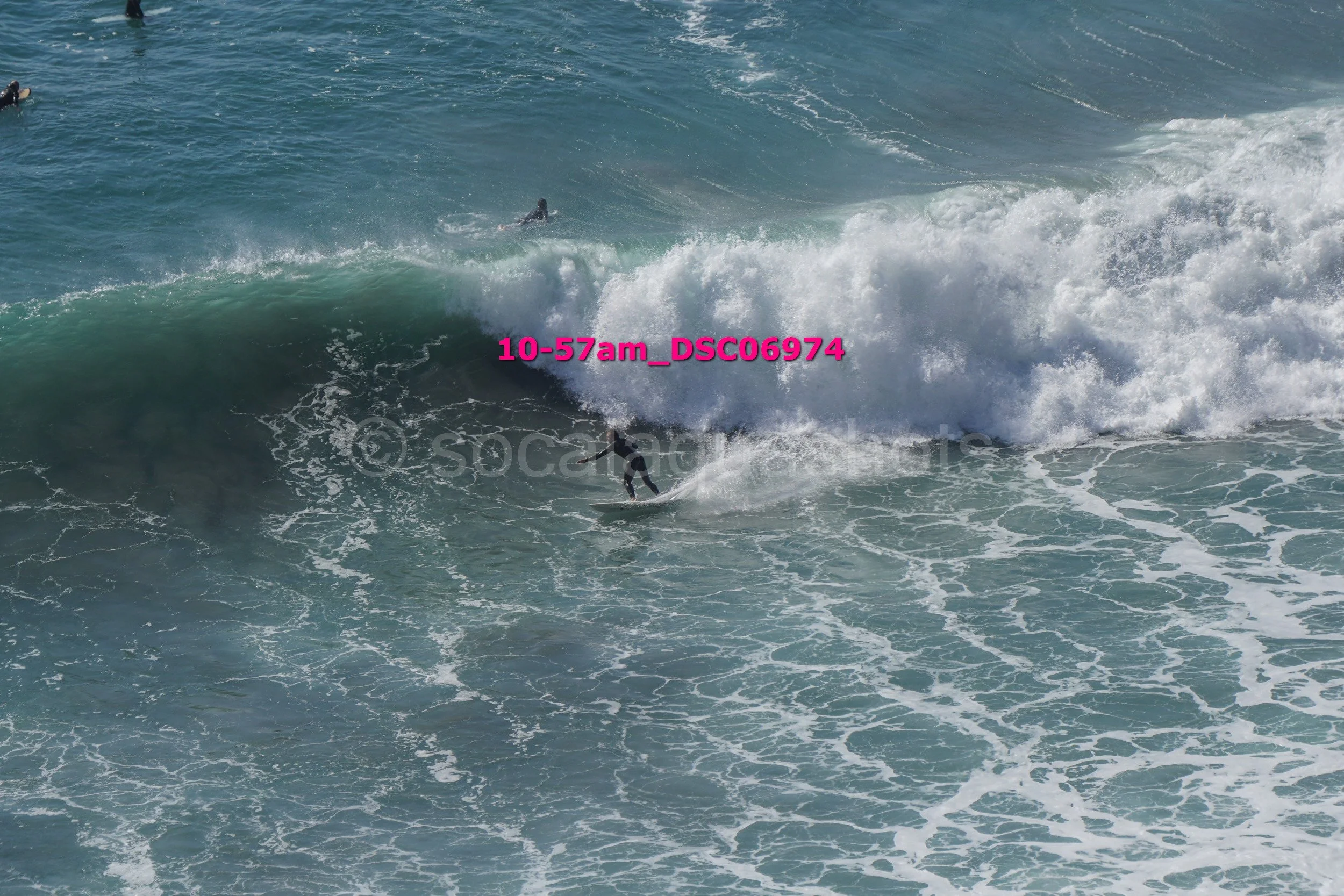 A surfer riding a large wave in the ocean, with other surfers visible in the water nearby.