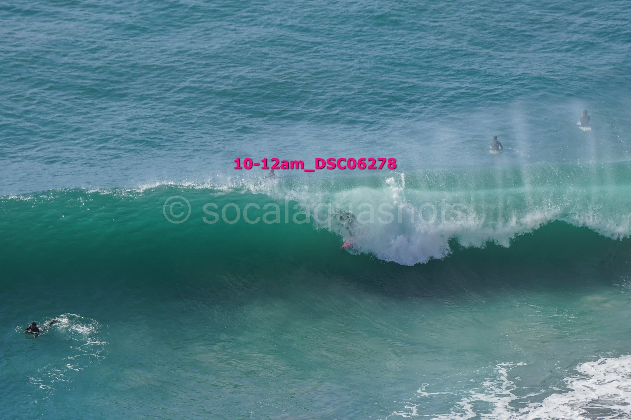 Surfers in the ocean near a large wave, some riding the wave and others waiting in the water.