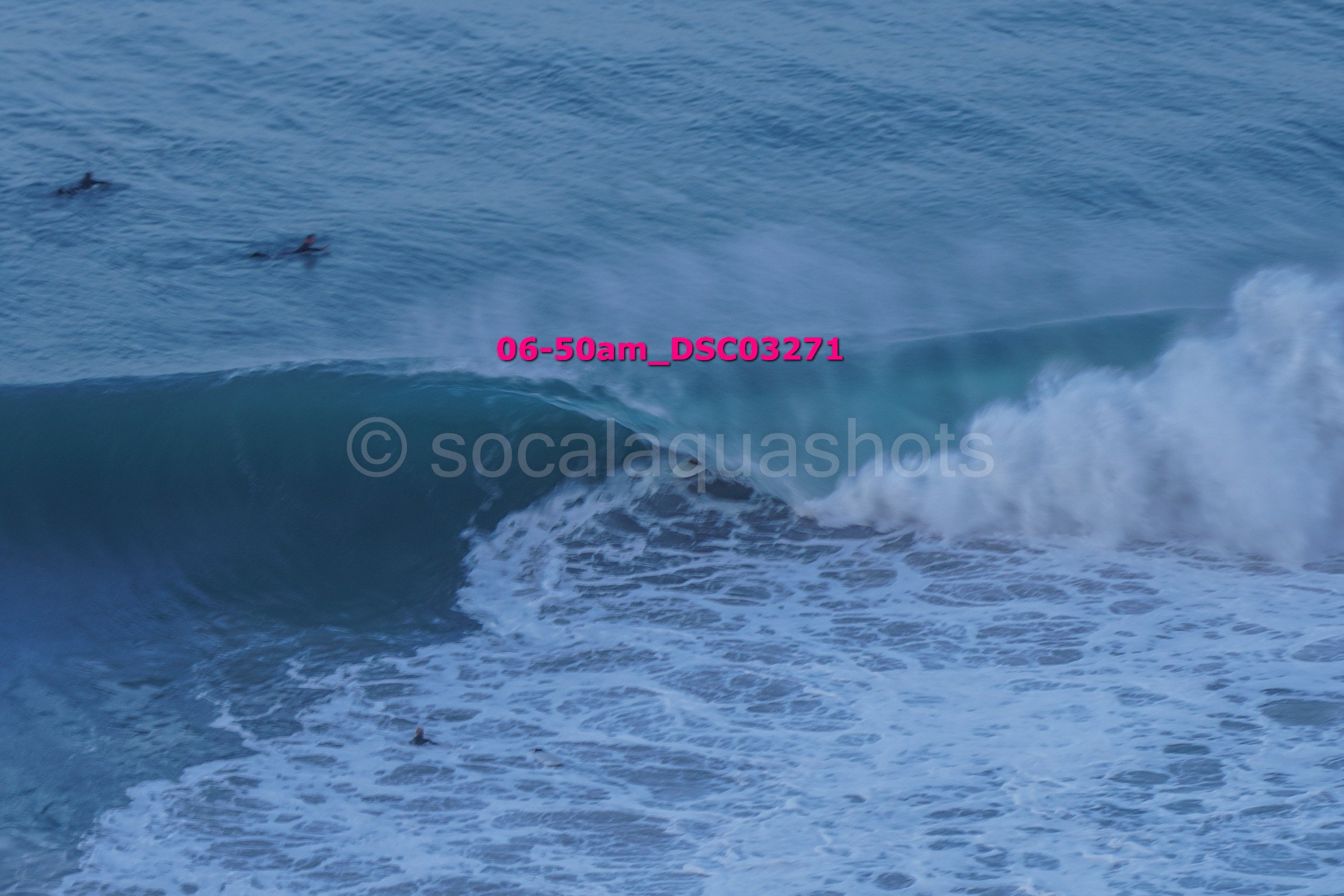 A large ocean wave with surfers in the water, one person riding the wave, and others swimming nearby.