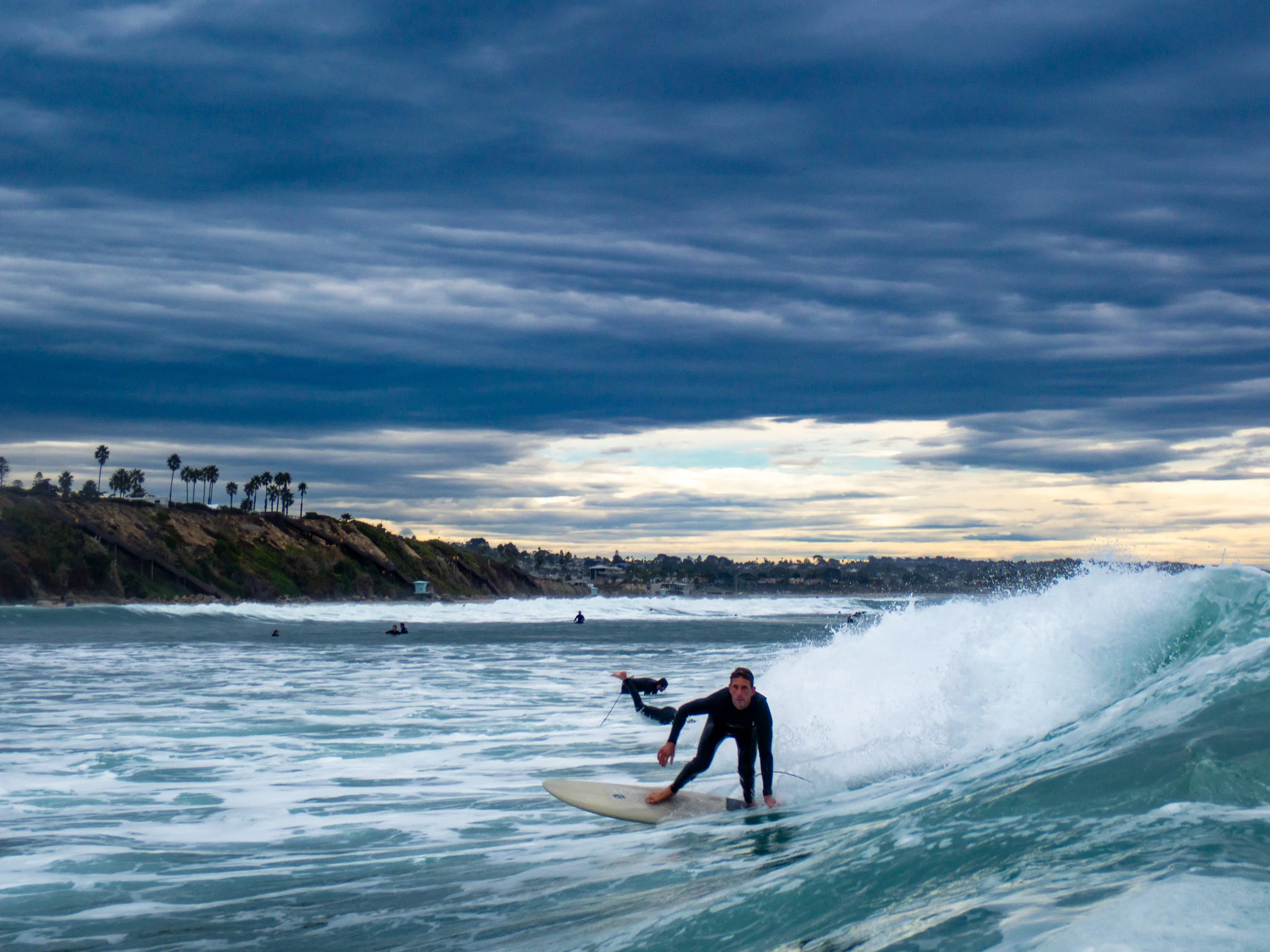 A person surfing on a wave in the ocean under a cloudy sky with a distant shoreline and palm trees in the background.