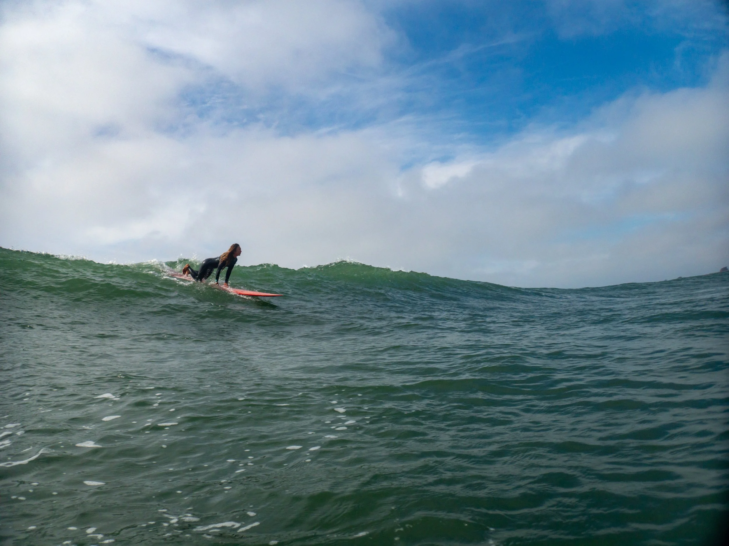 A person in a wetsuit surfboard riding a wave in the ocean under a partly cloudy sky.