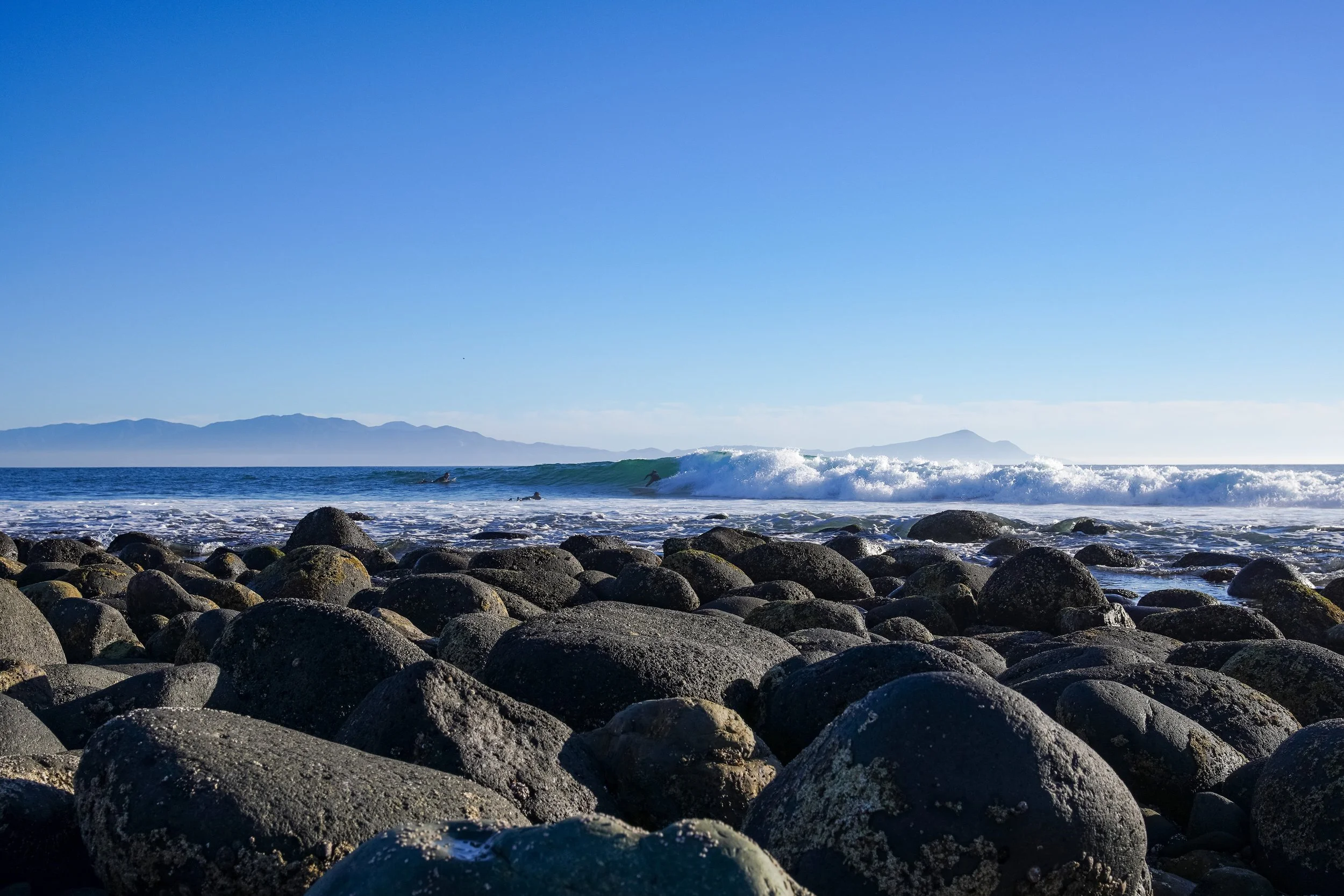A rocky beach with large dark stones in the foreground, ocean waves in the middle, and surfers riding the waves under a clear blue sky with distant mountains on the horizon.