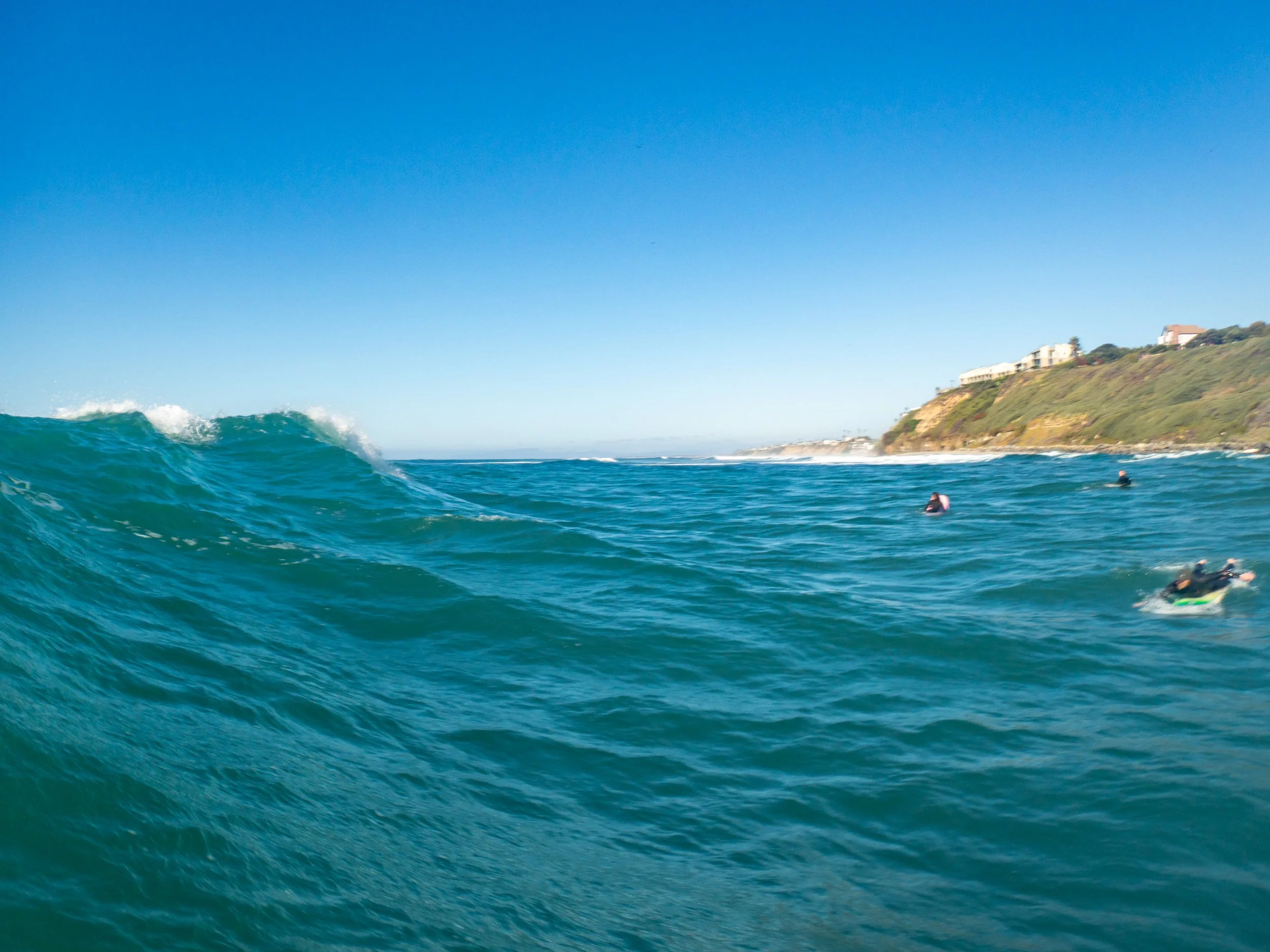 Ocean waves with surfers in the water near a coastal hillside with houses.