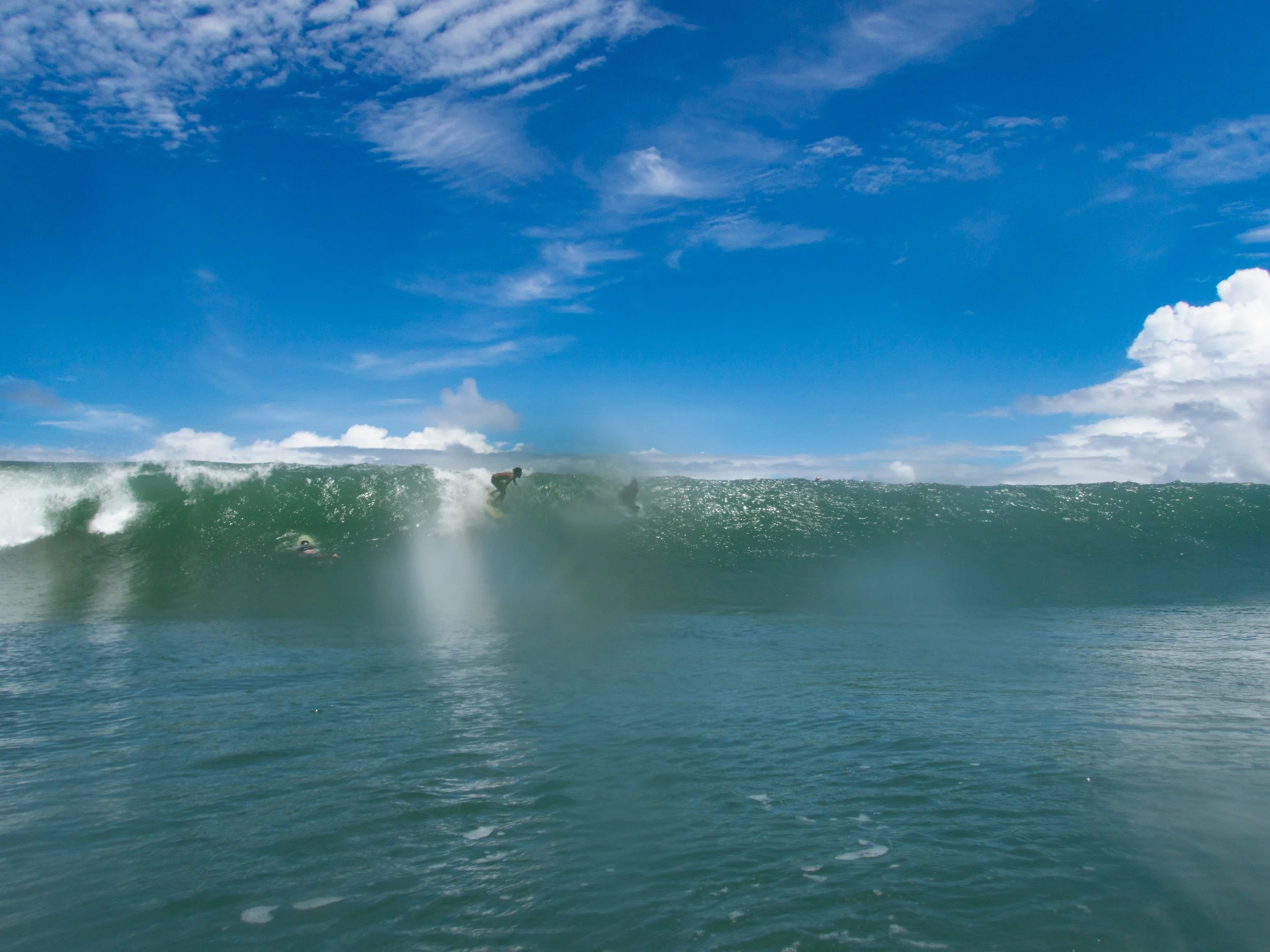 Surfers riding a large ocean wave under a clear blue sky.