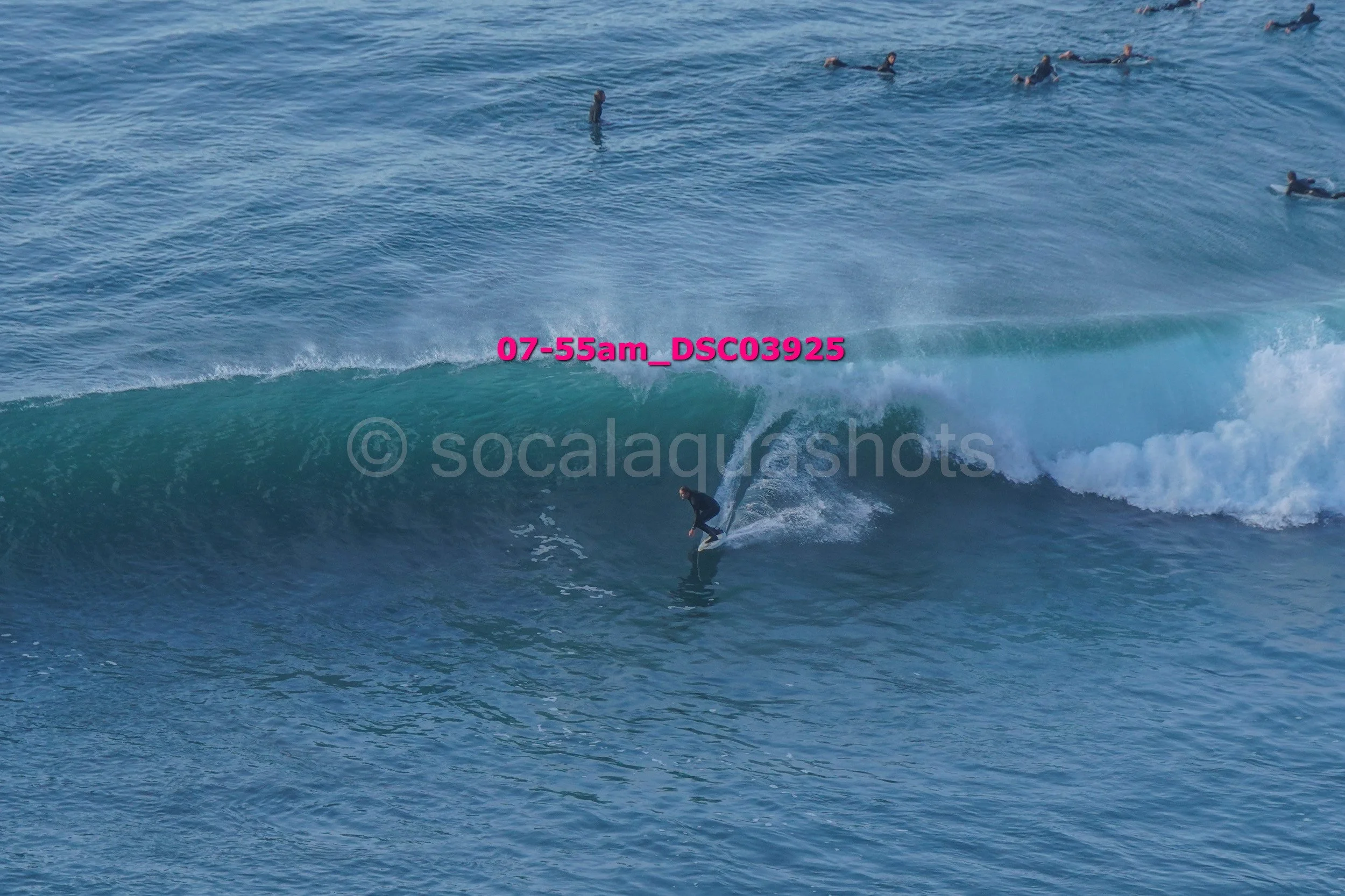 A person surfing on a wave in the ocean with several people swimming or floating in the water in the background.