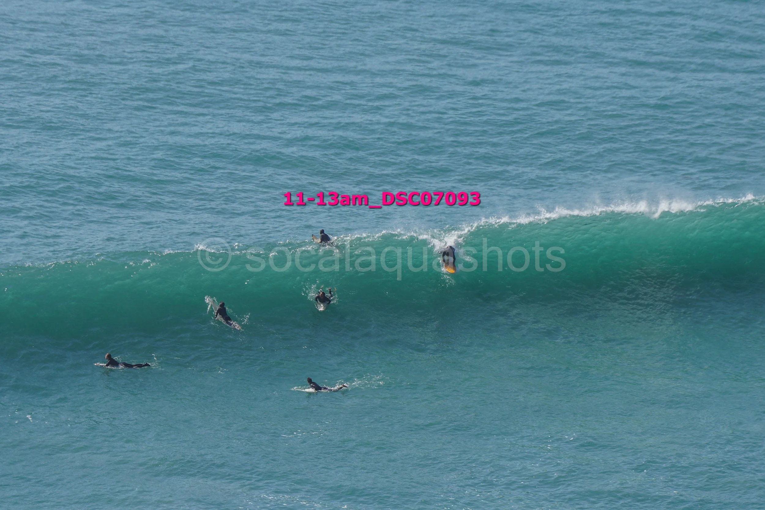 Several surfers riding and paddling on ocean waves, some on surfboards, in a large body of water under a blue sky.