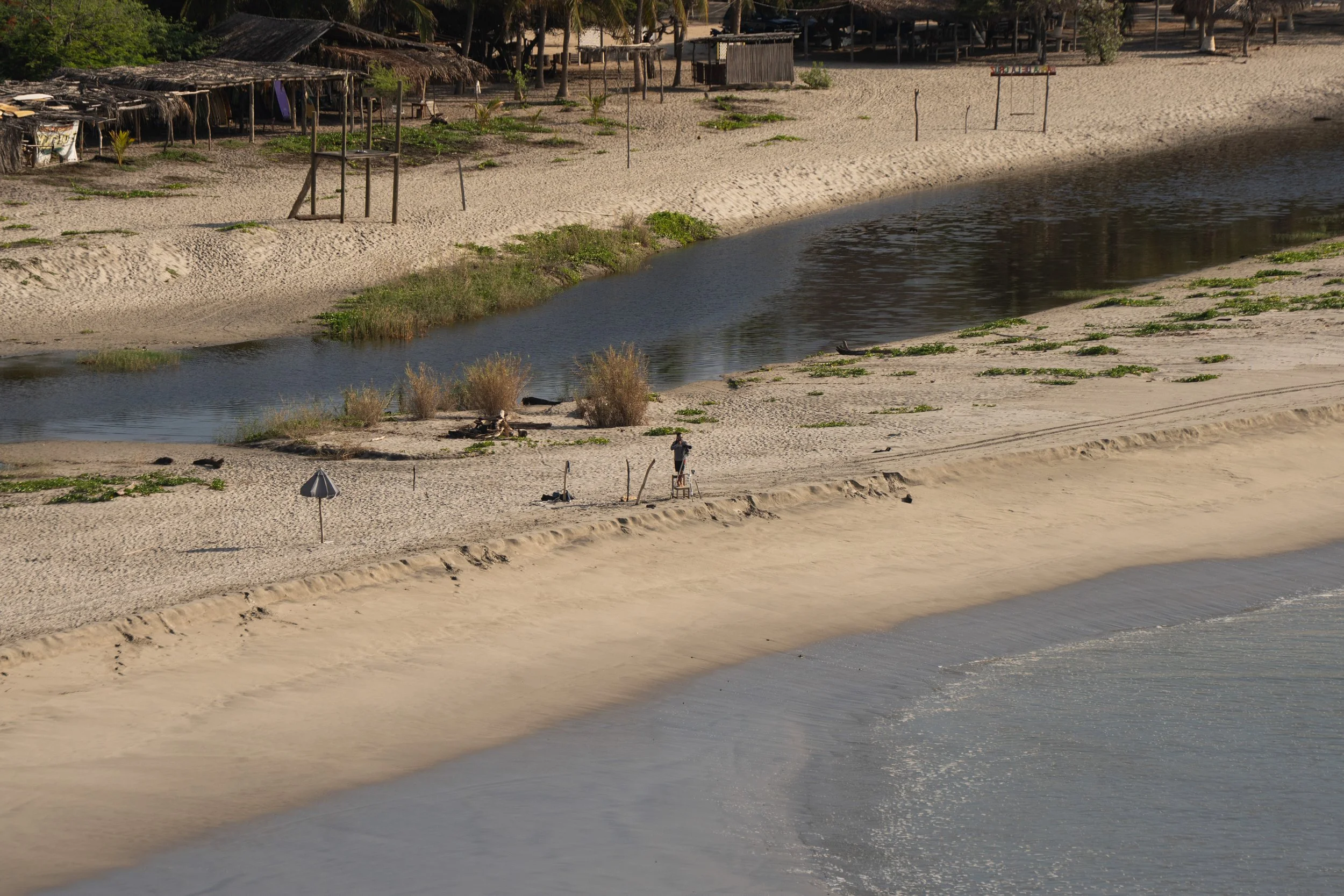 A beach scene with sandy shores, small huts with thatched roofs, a water body, and some scattered greenery. There are a few umbrellas and a person standing near the water.
