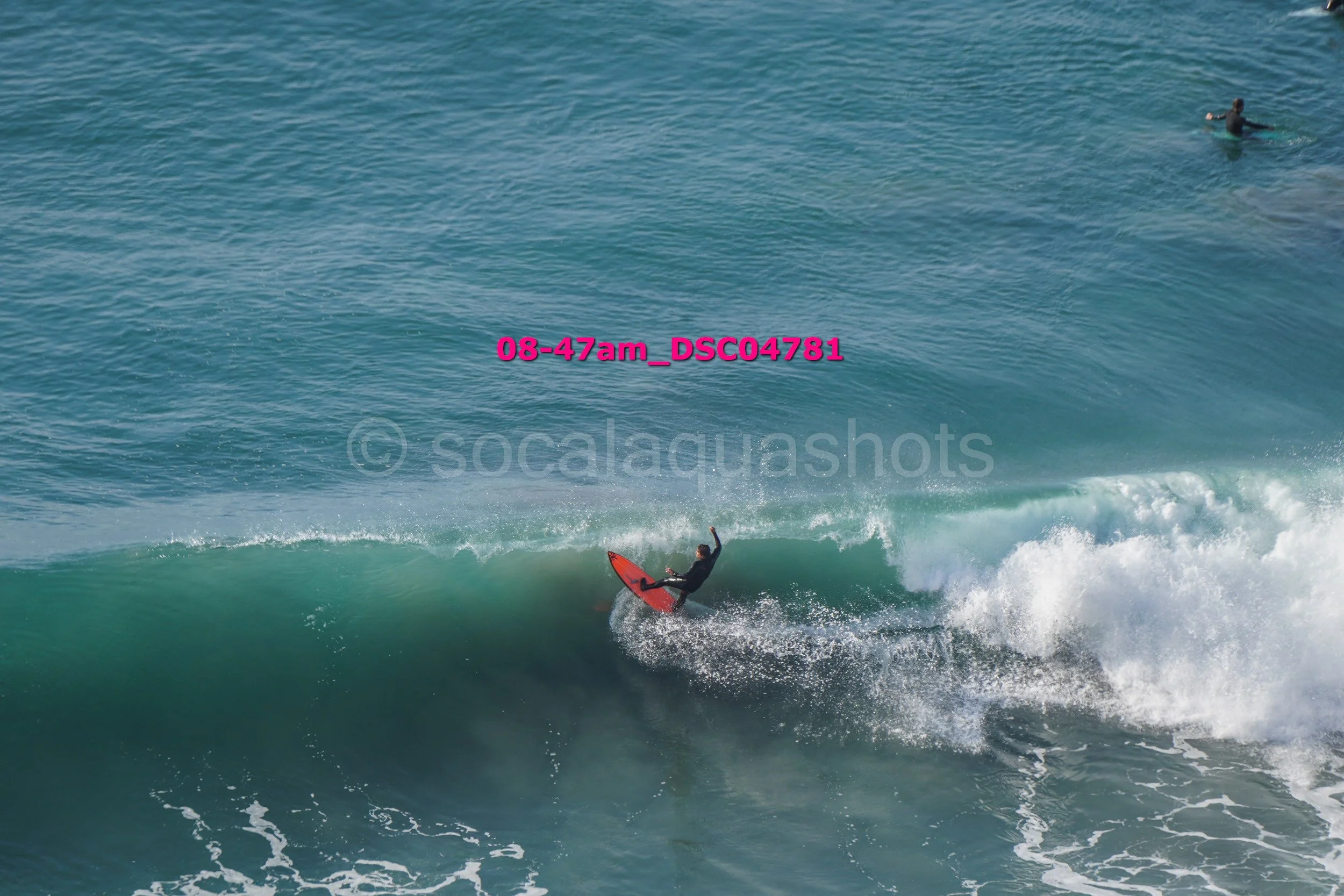 A person surfing on a red surfboard in the ocean with another person swimming in the background.