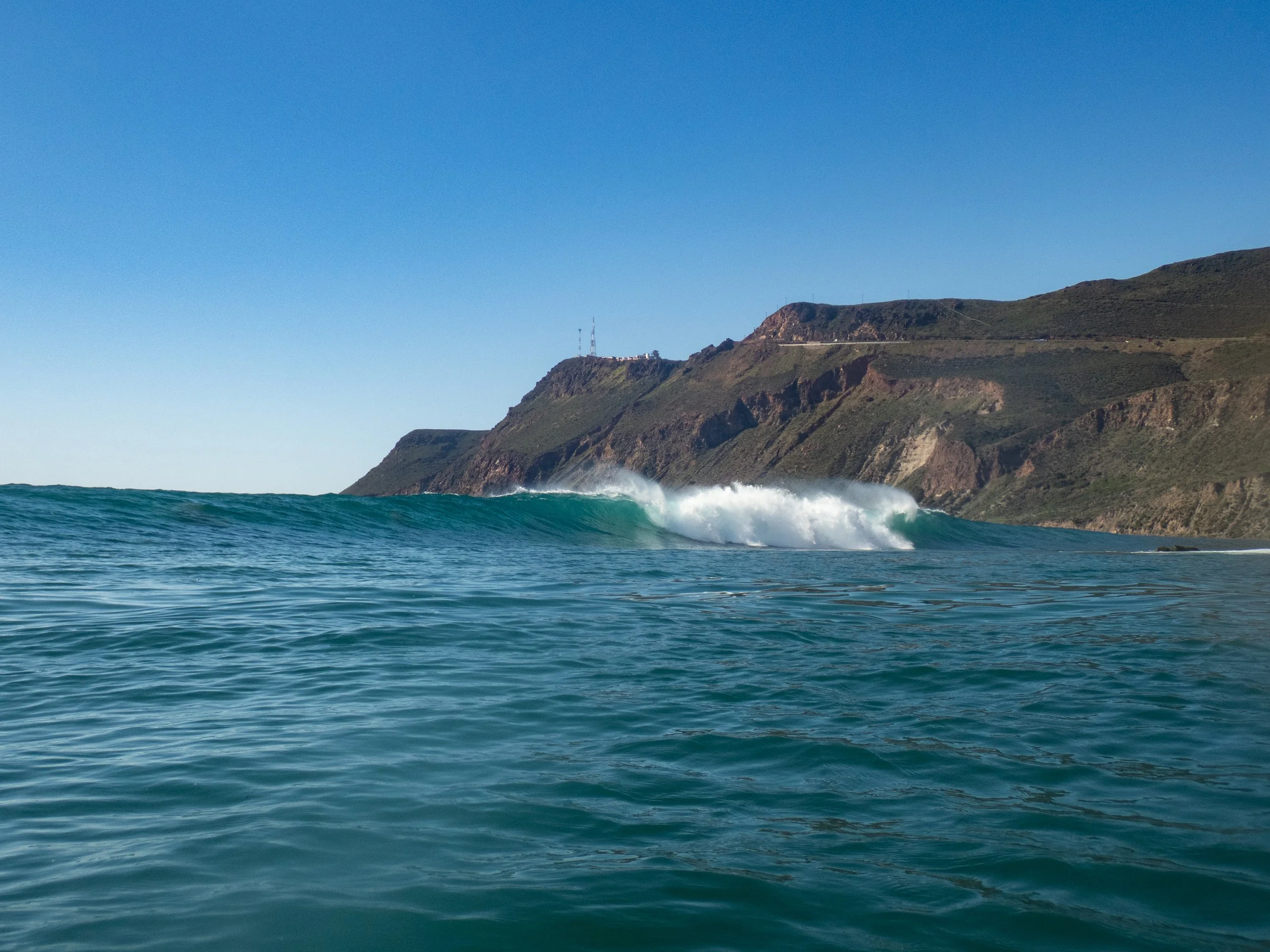 Ocean waves crashing near a rocky coastline with green cliffs and a clear blue sky.