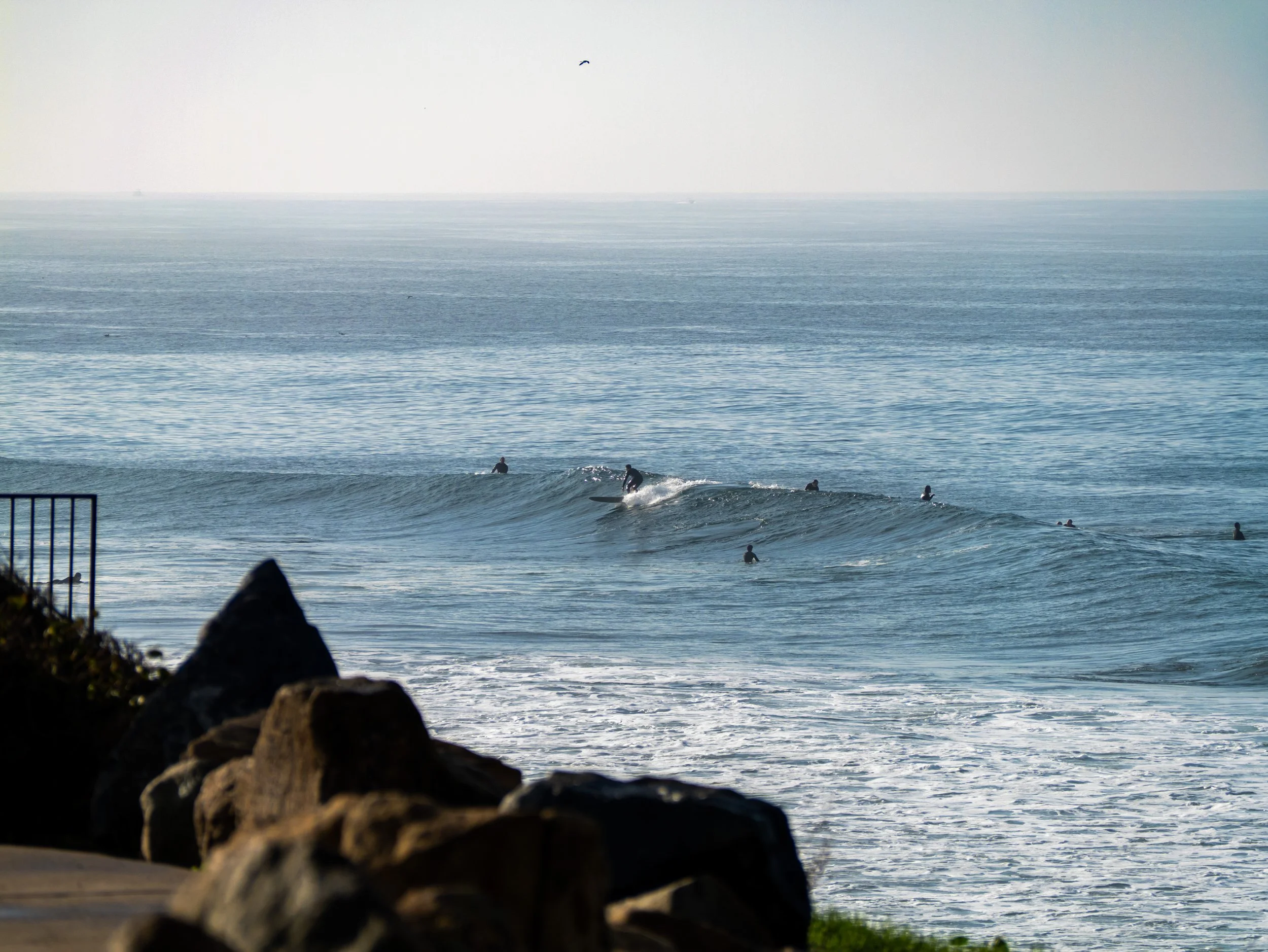 Surfers riding waves at the beach with rocks in the foreground and the sea extending to the horizon.