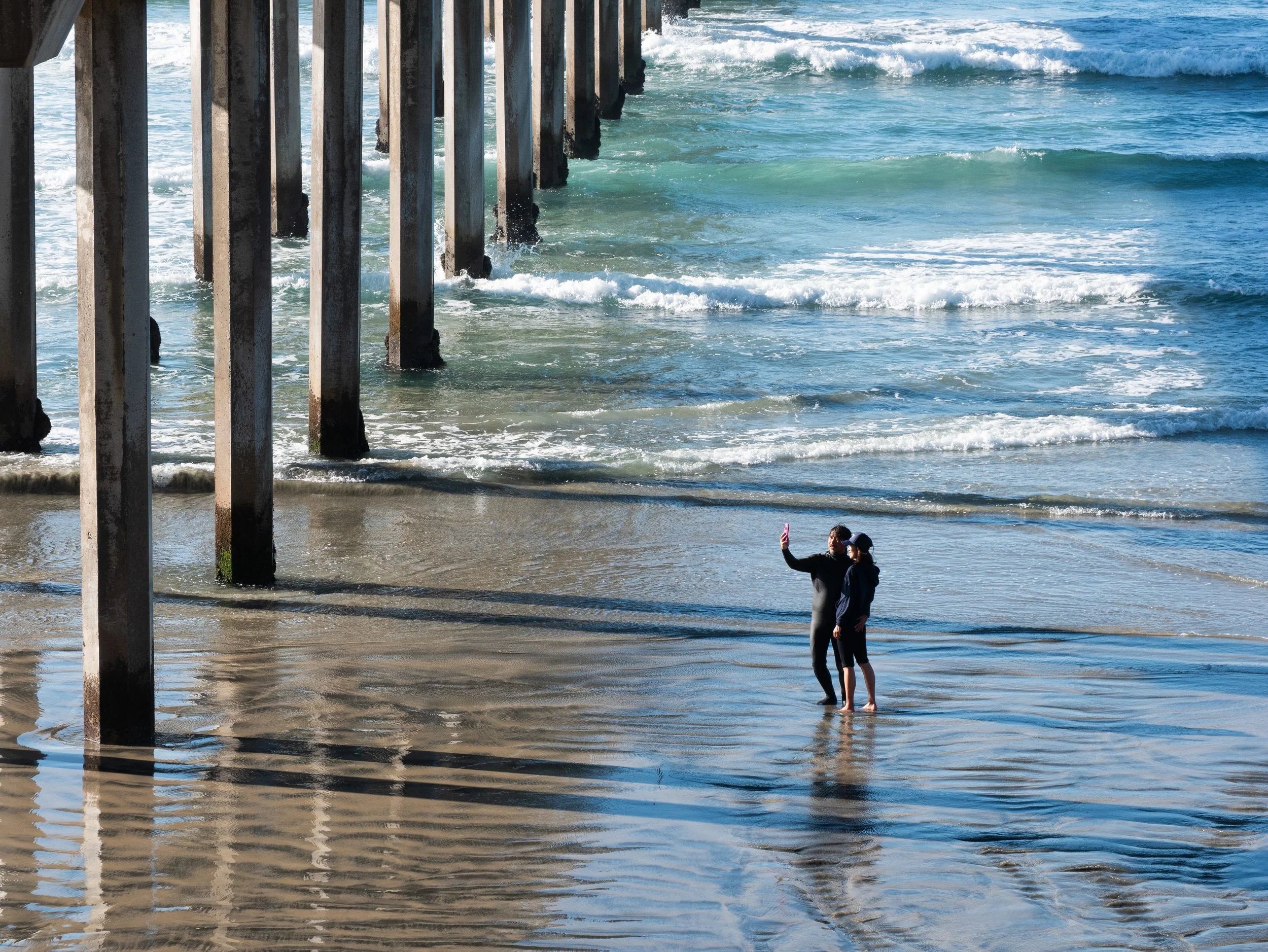 Two people taking a selfie on the beach near the ocean with a pier in the background.