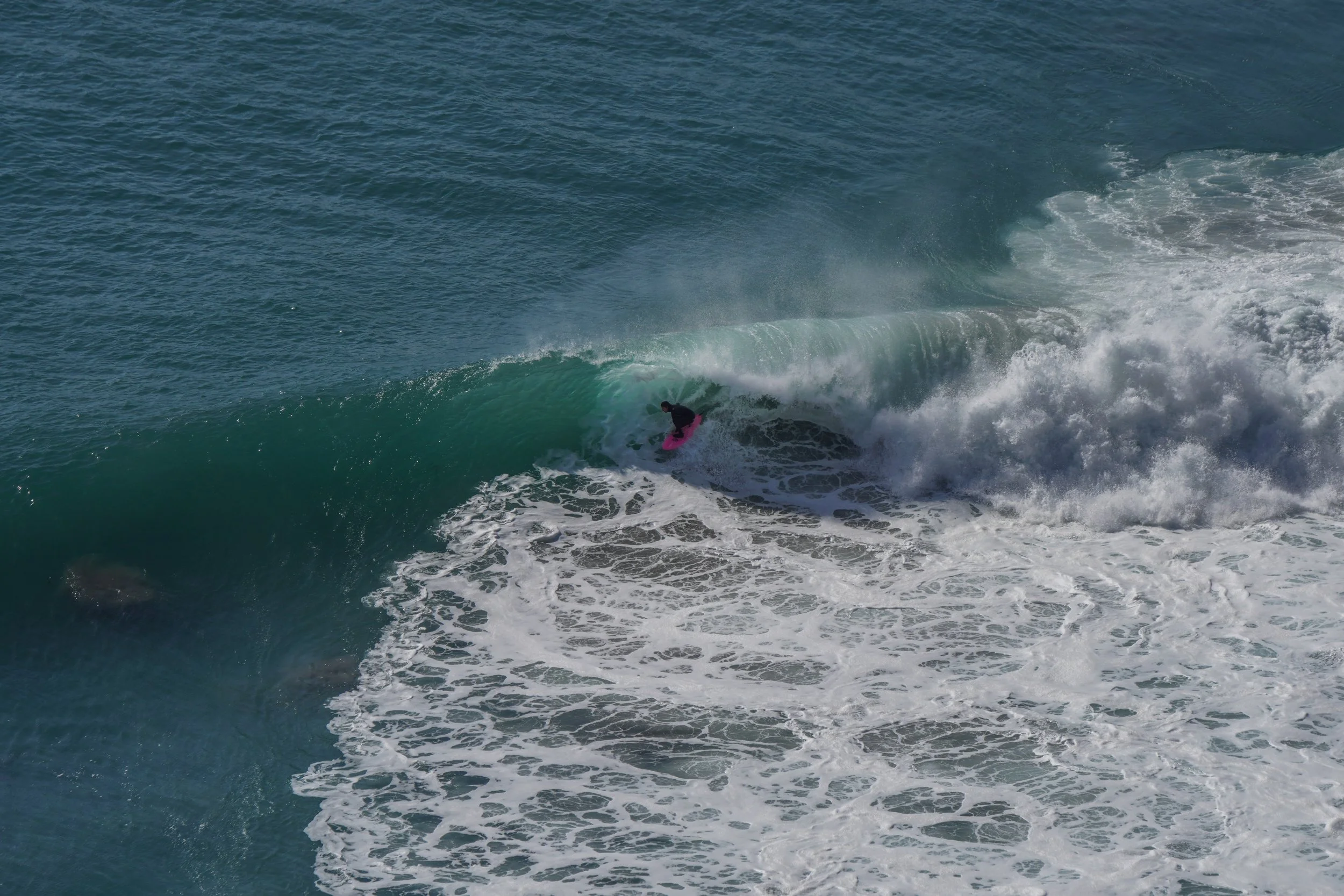 A surfer in black clothing riding inside a large, green wave with pink surfboard on a beach.