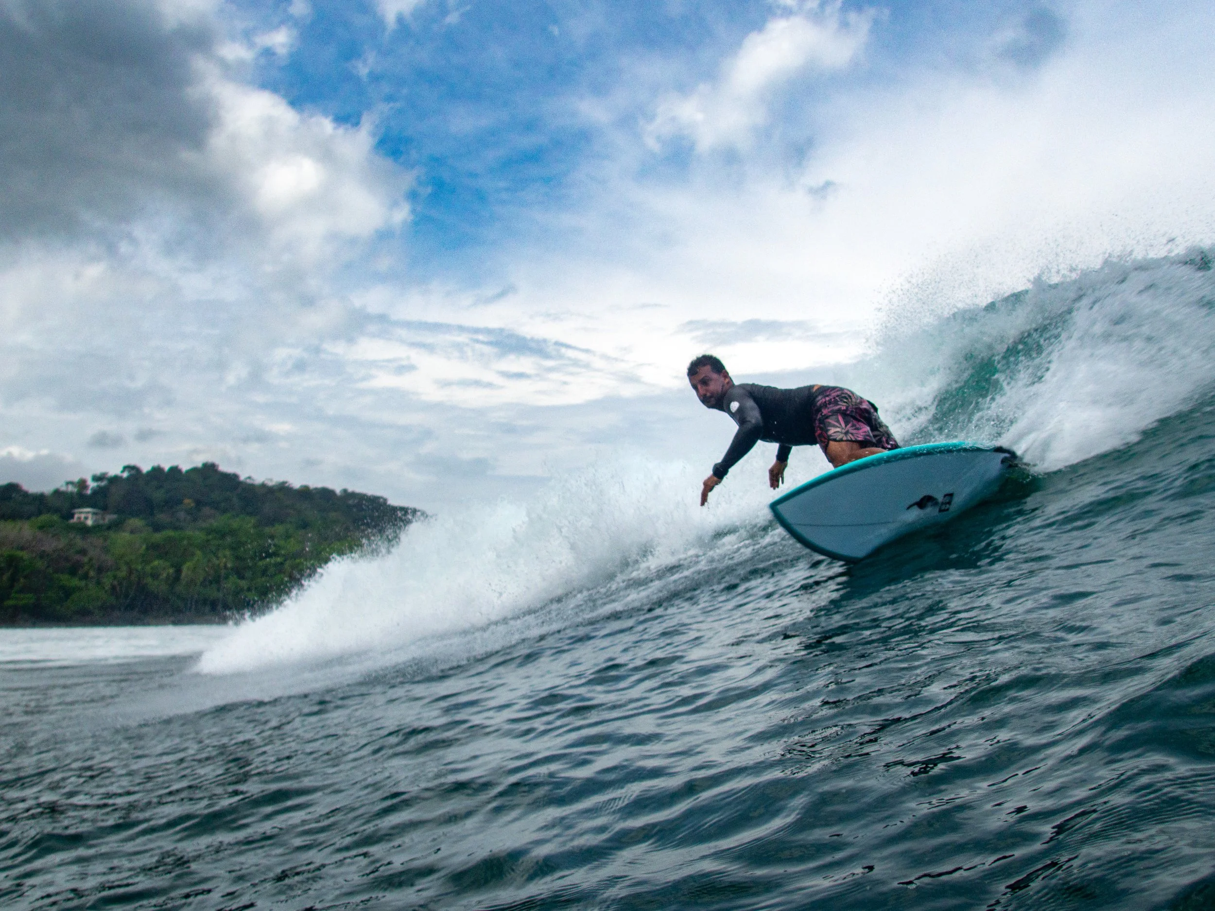 Surfer riding a wave in the ocean with a cloudy sky and greenery in the background.