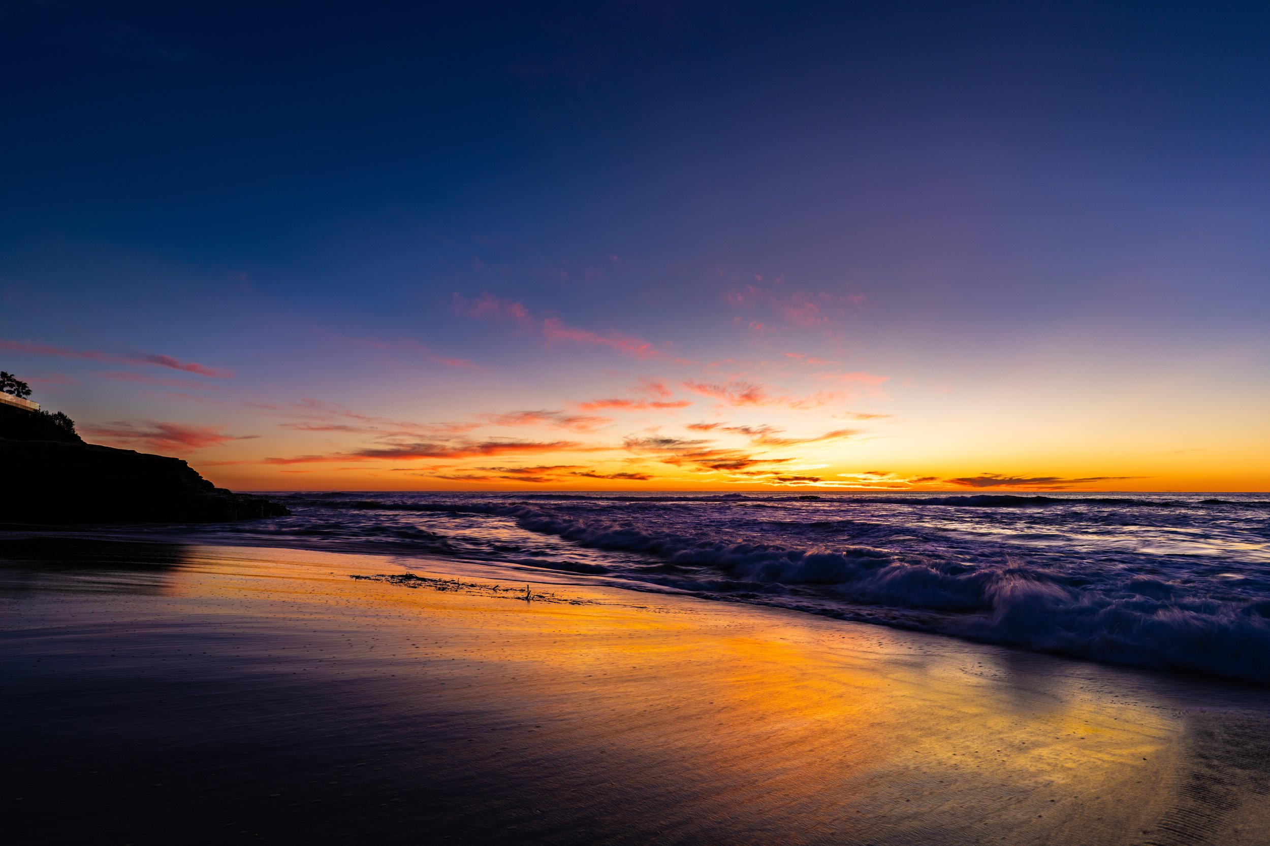 Sunset over the ocean with colorful sky, waves crashing on the shore, and a silhouette of a hillside on the left.