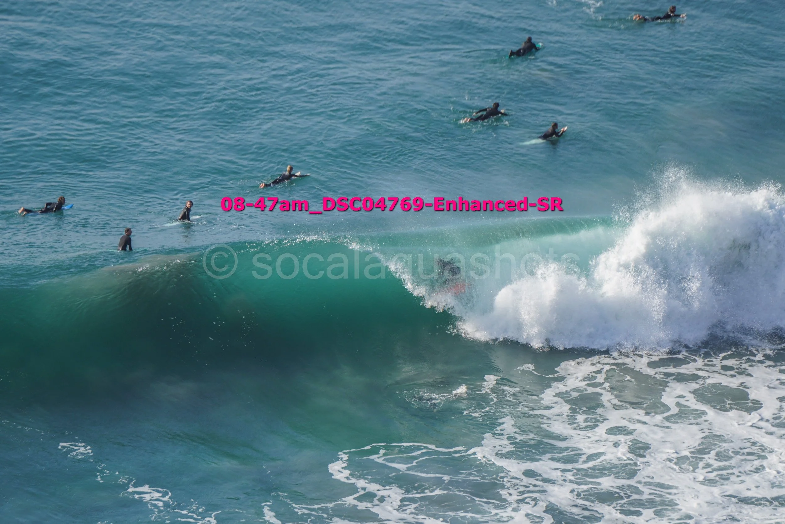Multiple surfers in wetsuits swimming and waiting in the ocean, with one surfer riding a large wave.