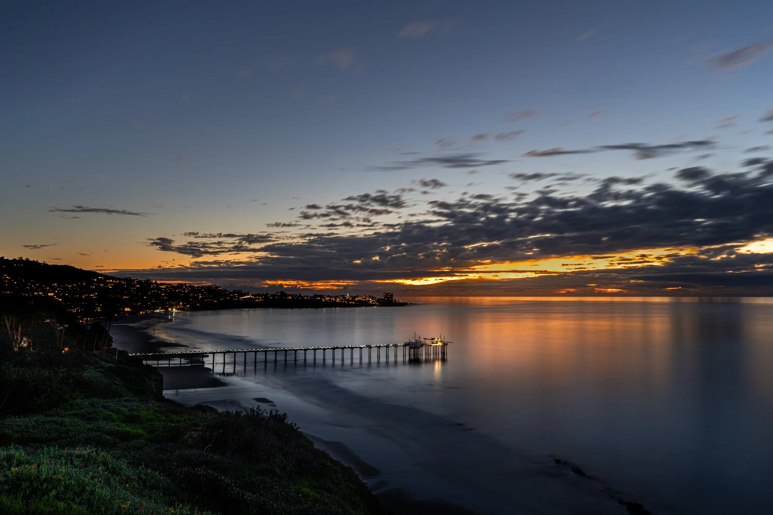 Sunset over a calm body of water with a pier extending into the water, a shoreline with houses and a hill on the left side, partially cloudy sky with orange and yellow hues.