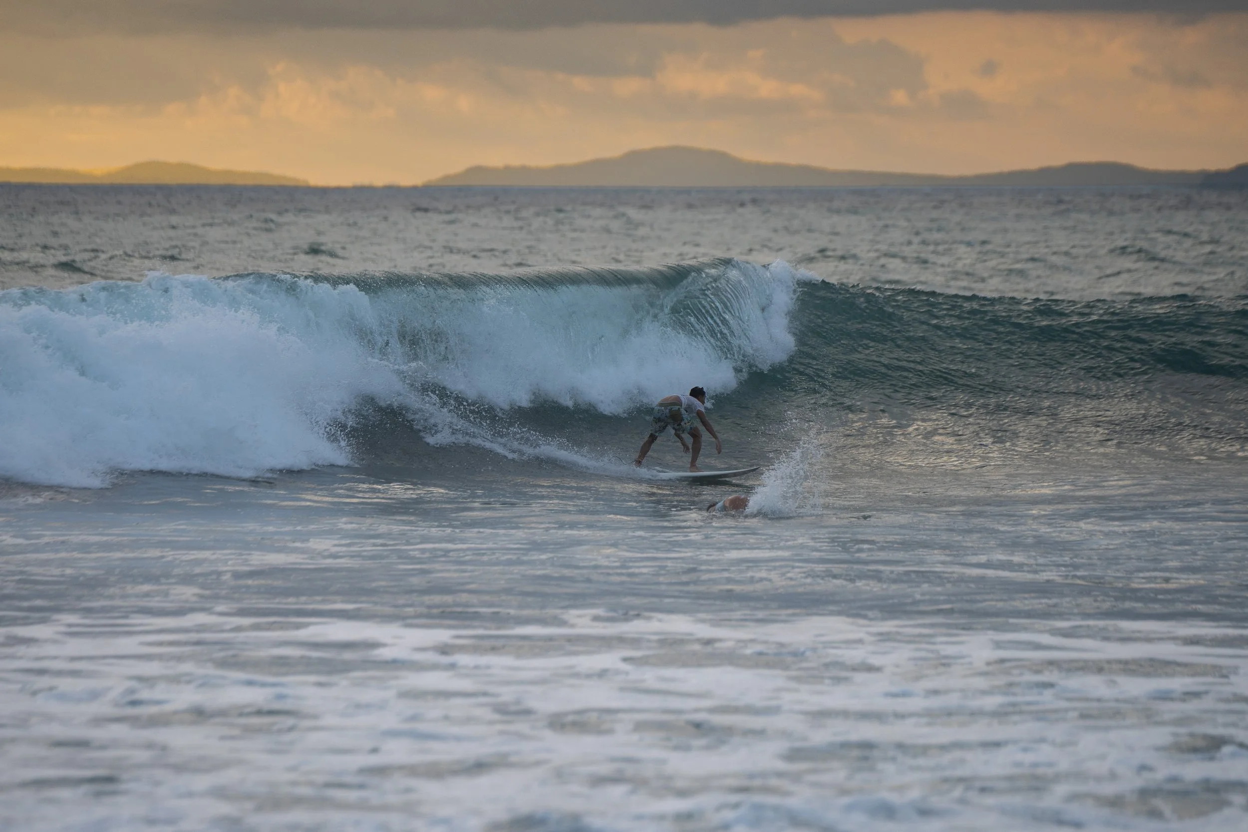 A person surfing on a wave in the ocean during sunset, with distant islands visible in the background.