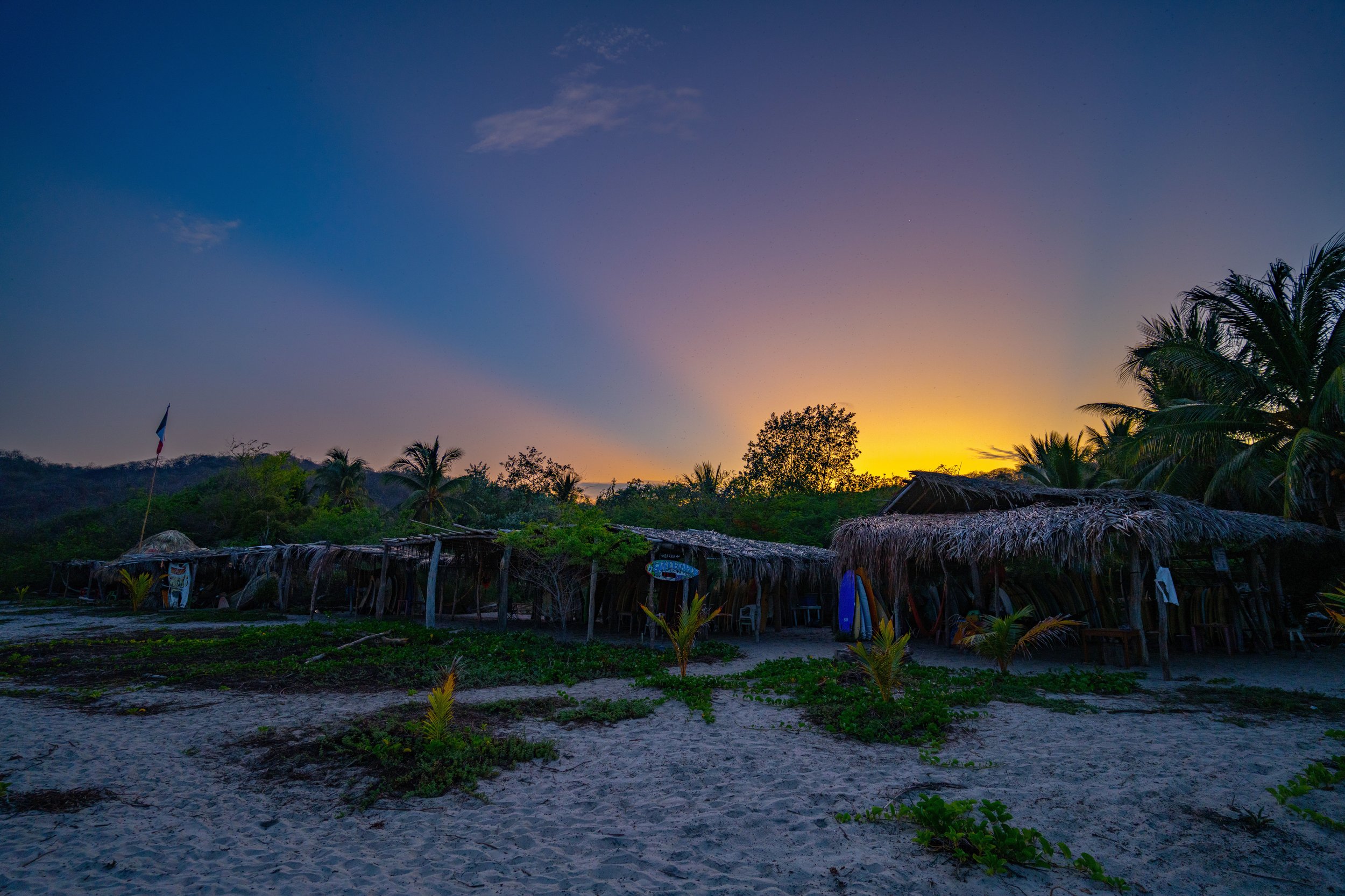 Sunset over a tropical village with thatched roofs, palm trees, and a sandy area in the foreground.
