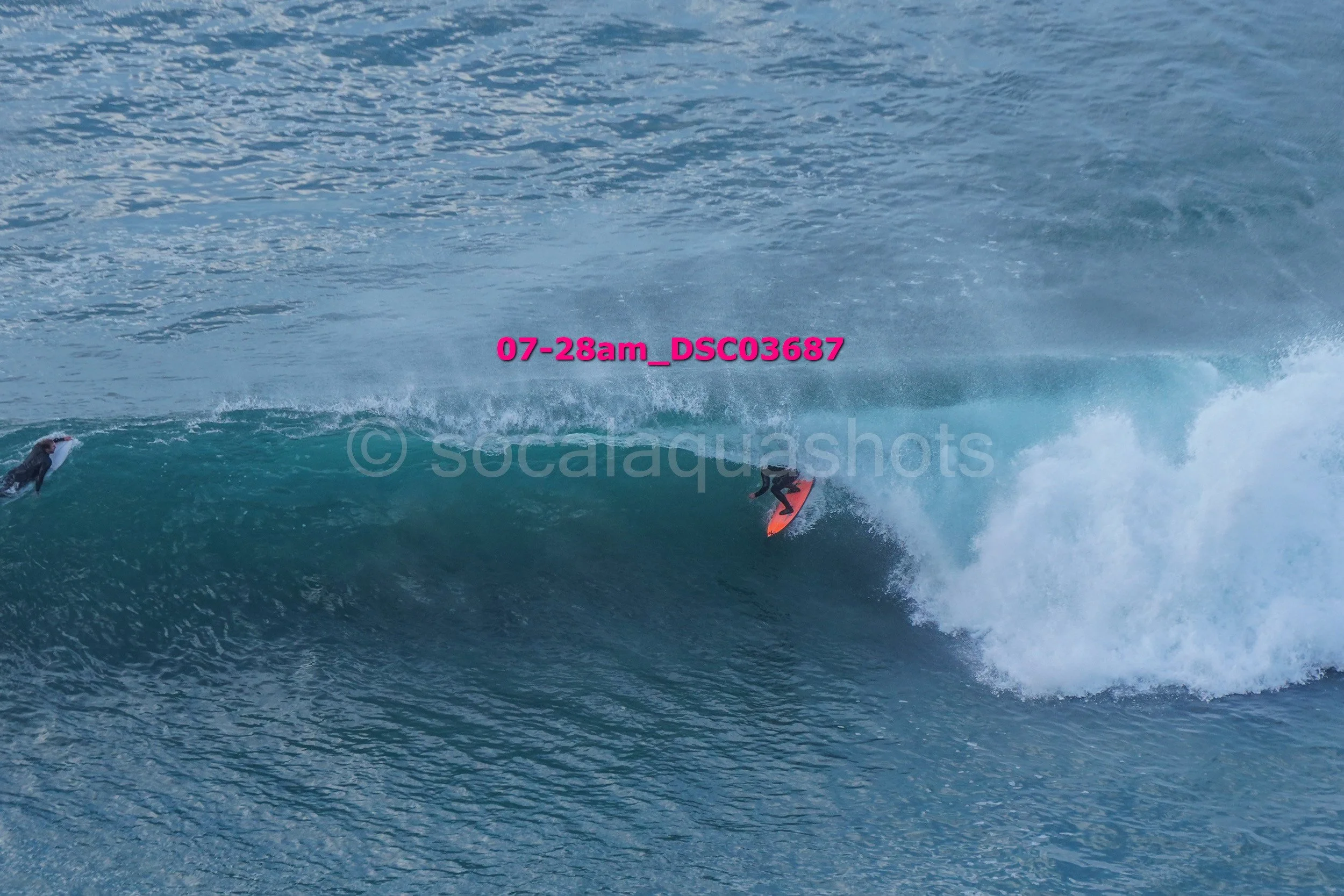 Surfer riding a large wave with another surfer in the background, ocean waters, daytime.