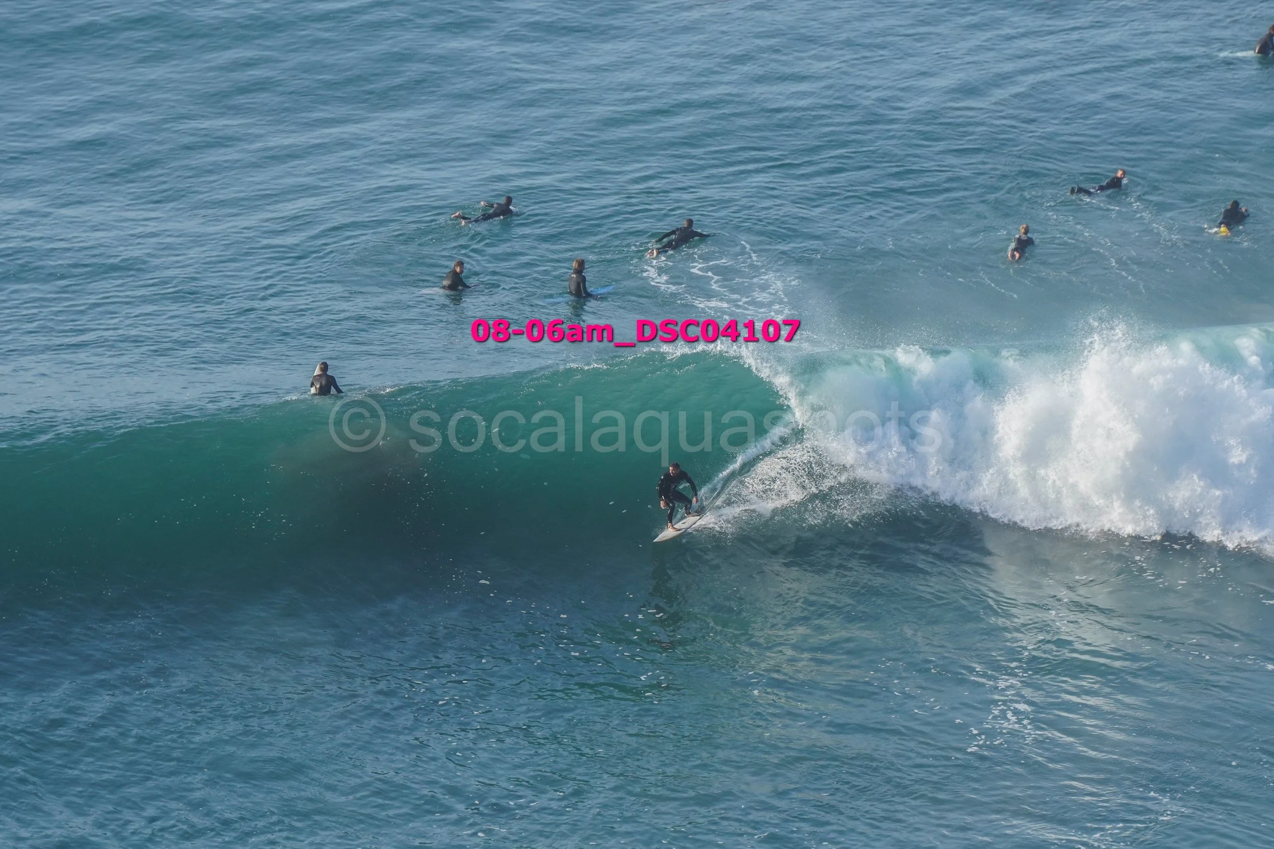 A person surfing on a large wave with several surfers waiting in the water in the background.