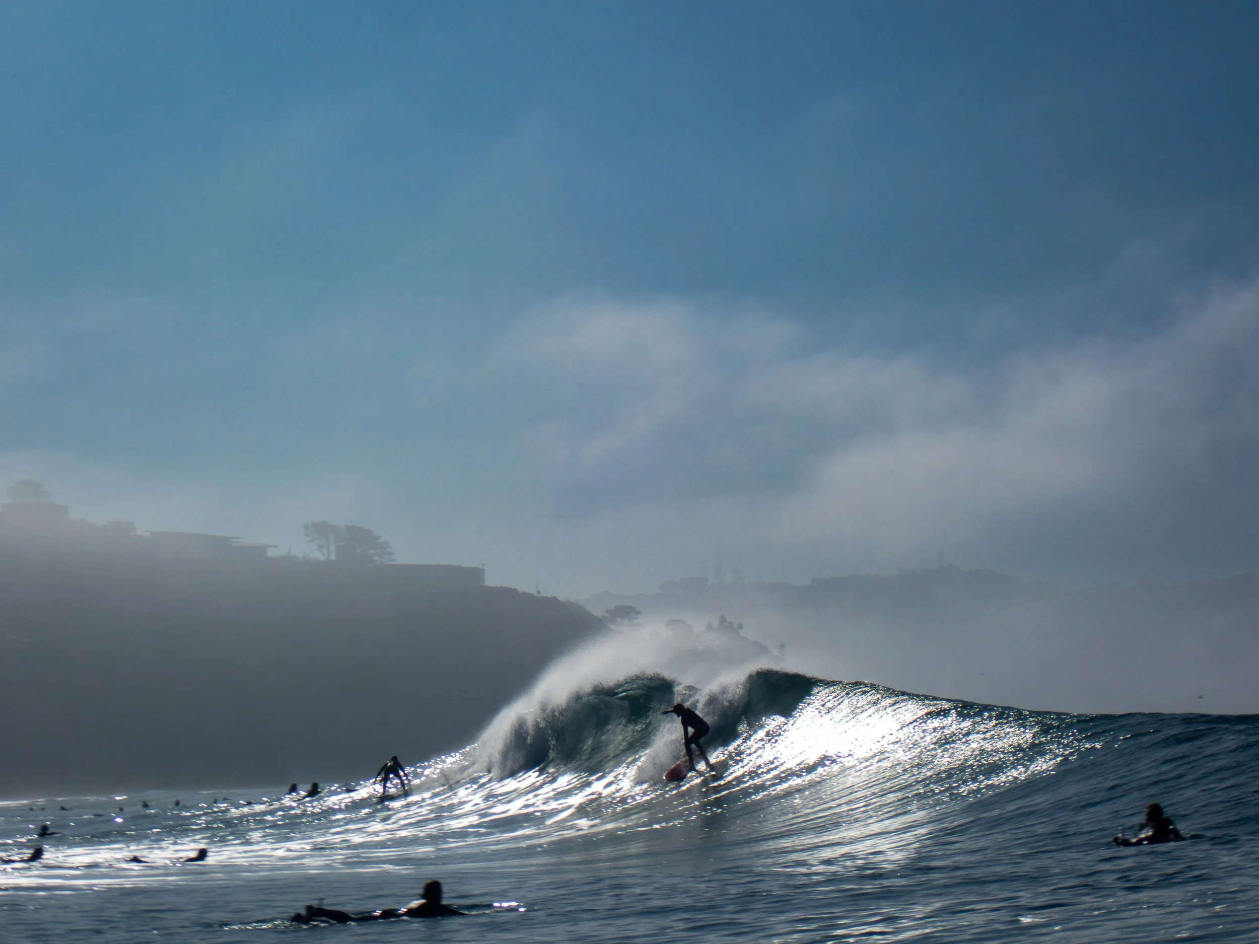 Surfer riding a wave in the ocean, others floating on surfboards, with a foggy shoreline in the background under a blue sky.