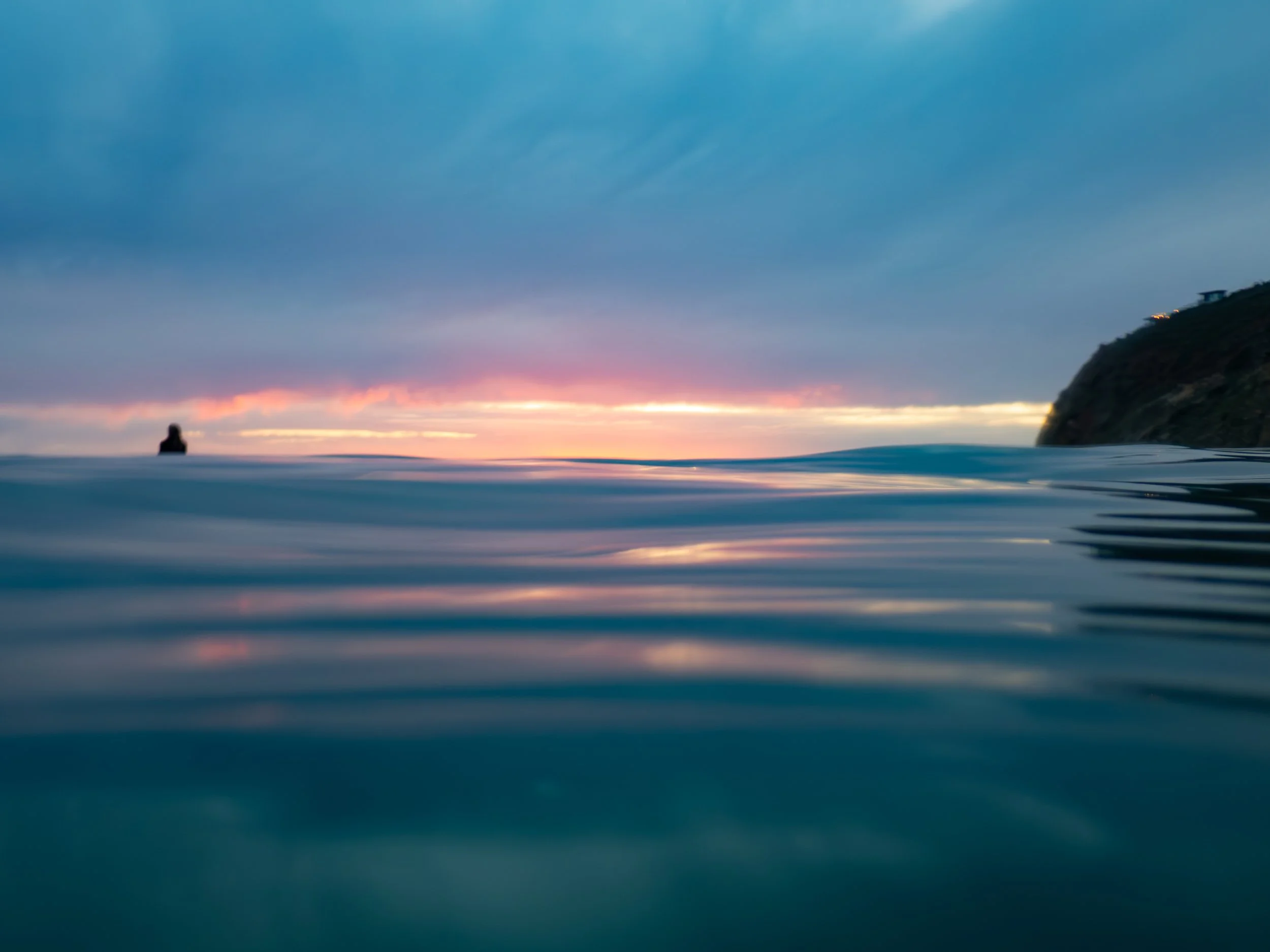 Ocean scene at sunset with a lone swimmer, a distant shoreline, and a cloudy sky.