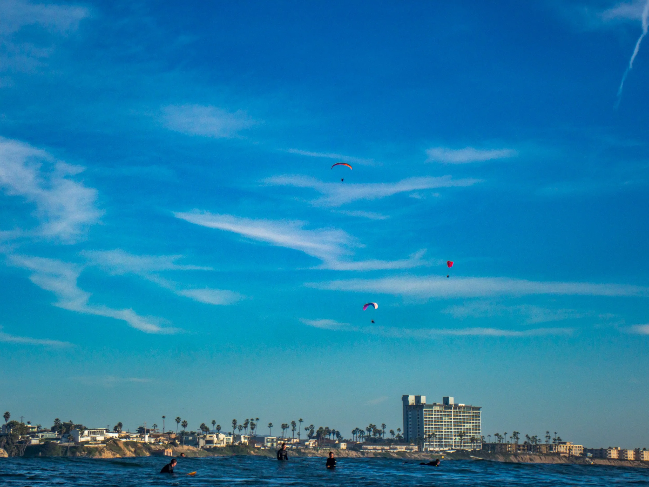 People surfing and parasailing over the ocean with a cityscape and palm trees in the background under a blue sky with wispy clouds.