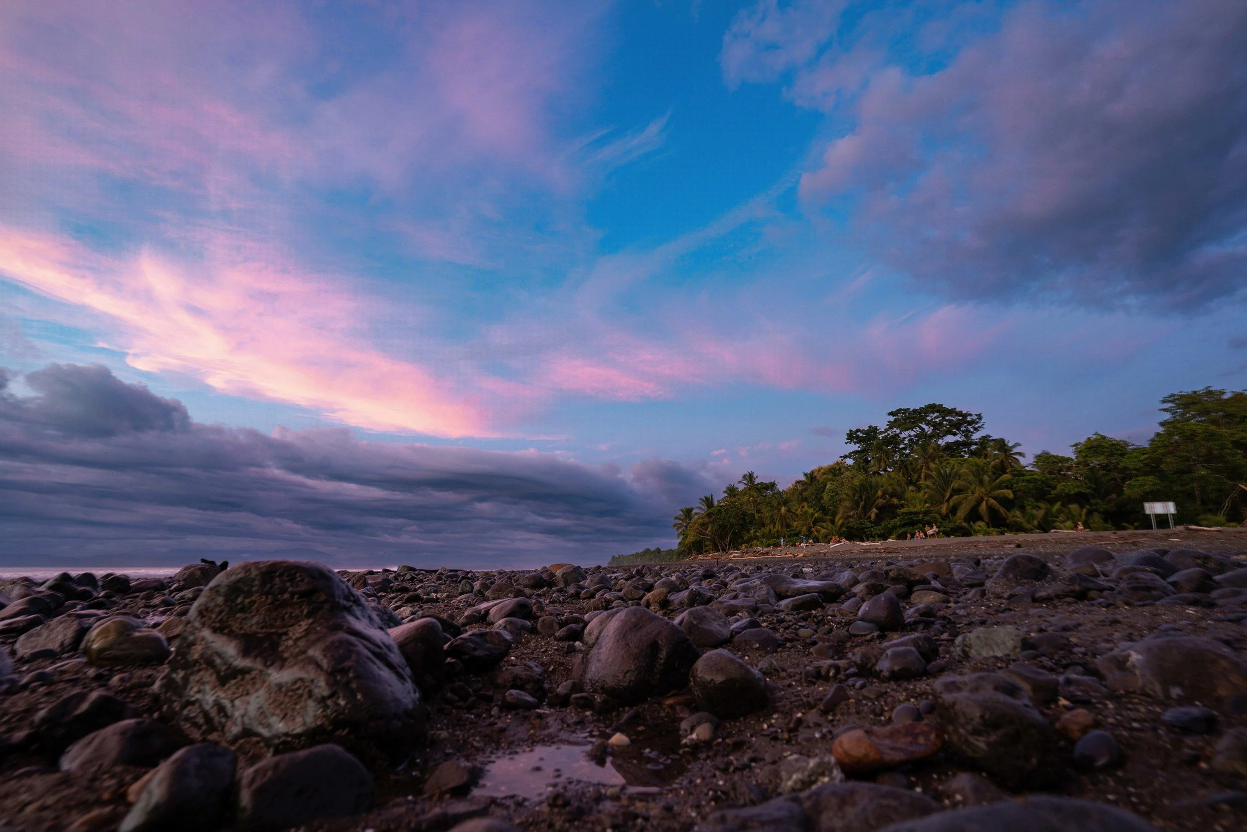 A rocky beach at sunset with a line of trees in the distance, colorful sky with pink and purple clouds.