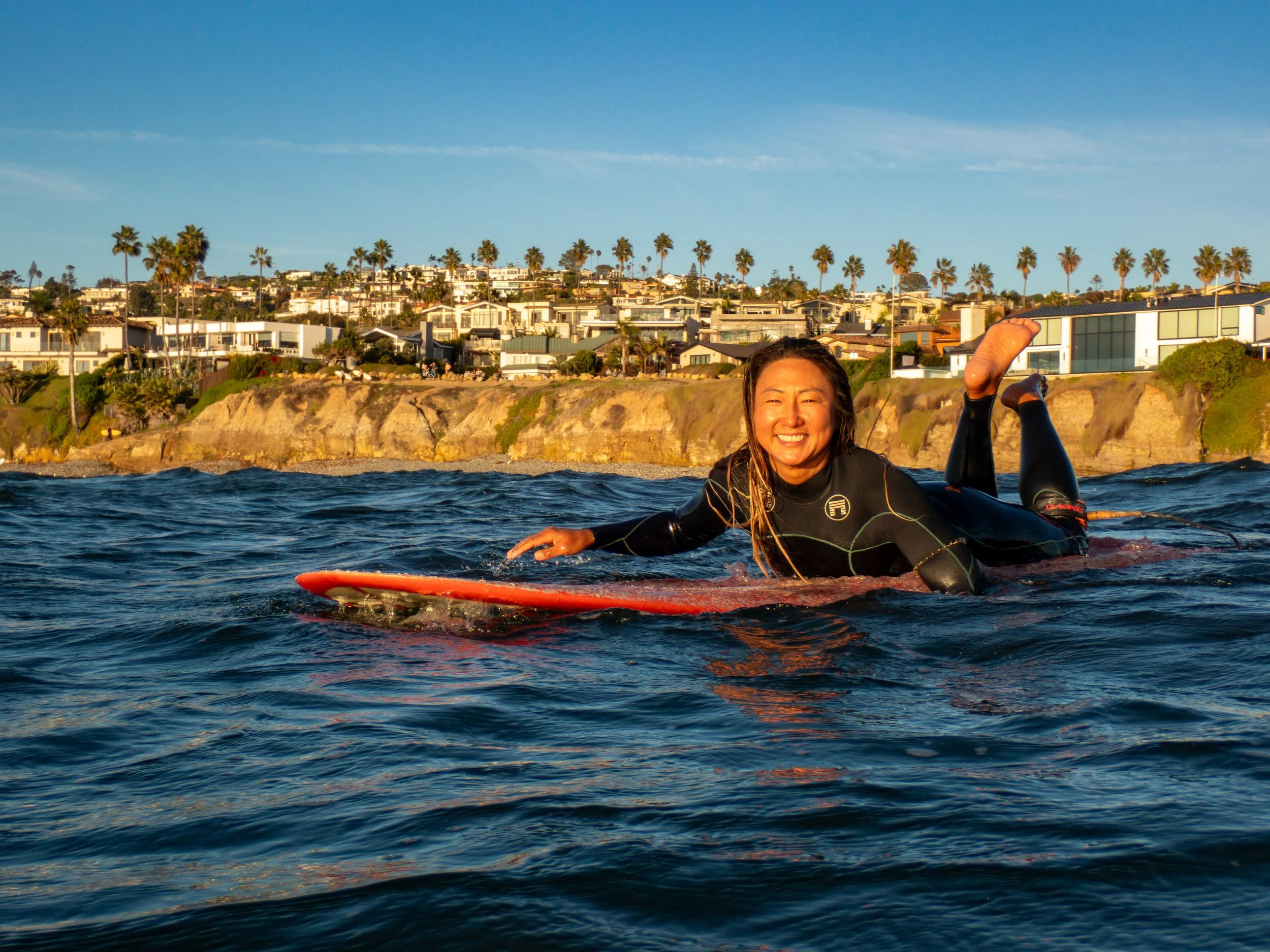 Woman in a wetsuit smiling on her surfboard in the water near the beach with houses and palm trees in the background.