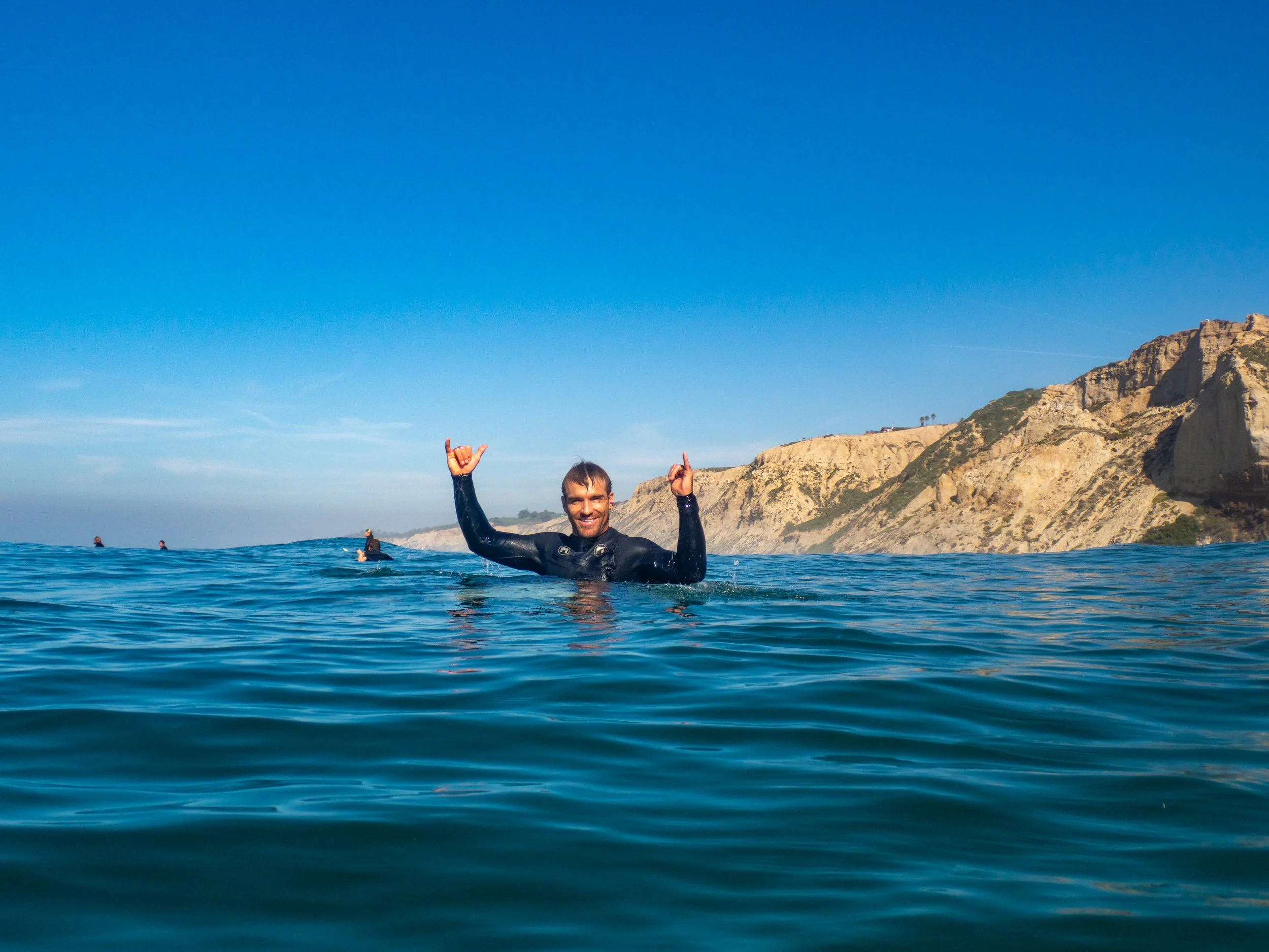 A man in a wetsuit smiling and making a hand sign while swimming in the ocean with cliffs in the background.