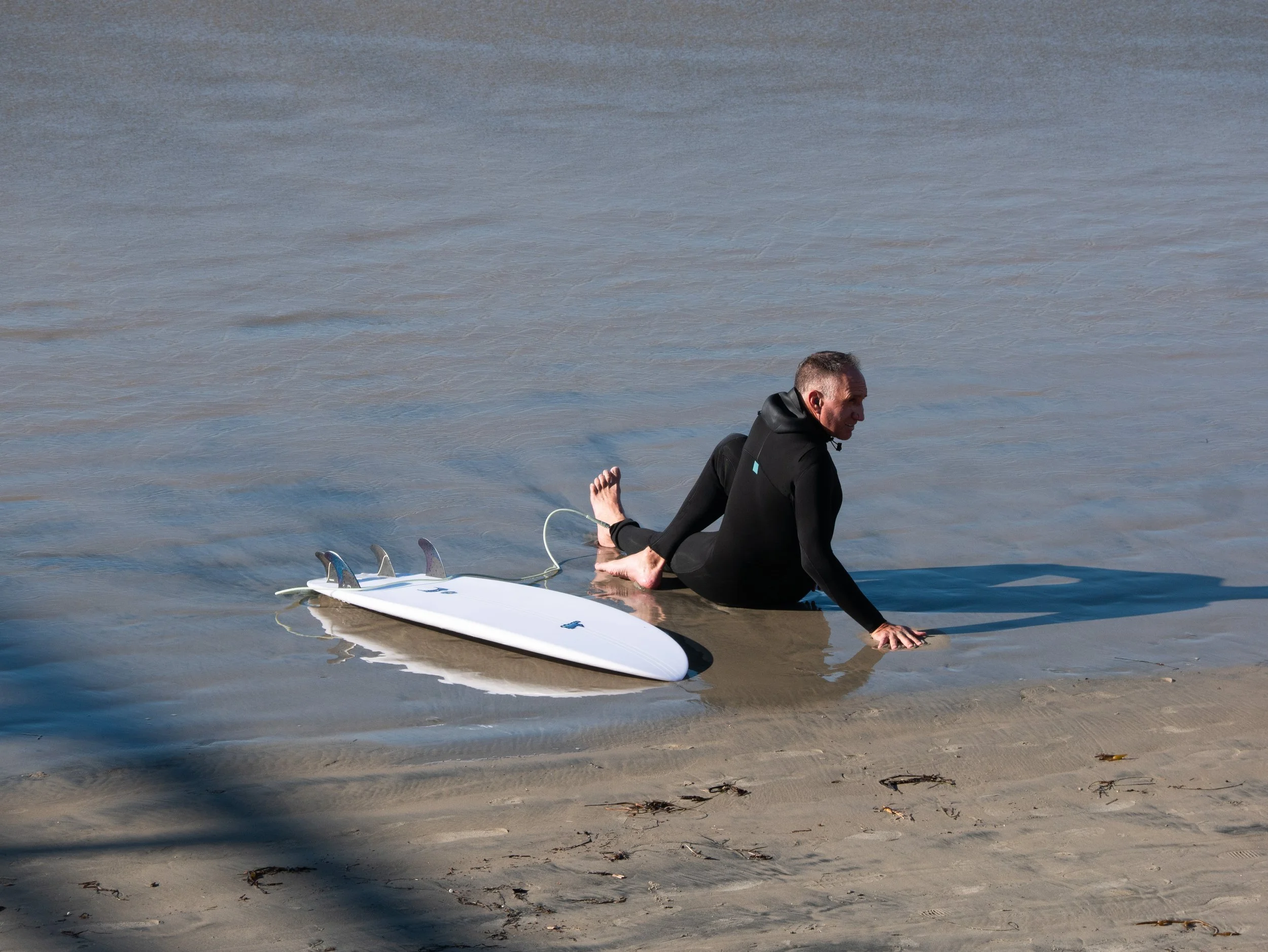 A man in a black wetsuit sitting on the beach next to a surfboard in shallow water.