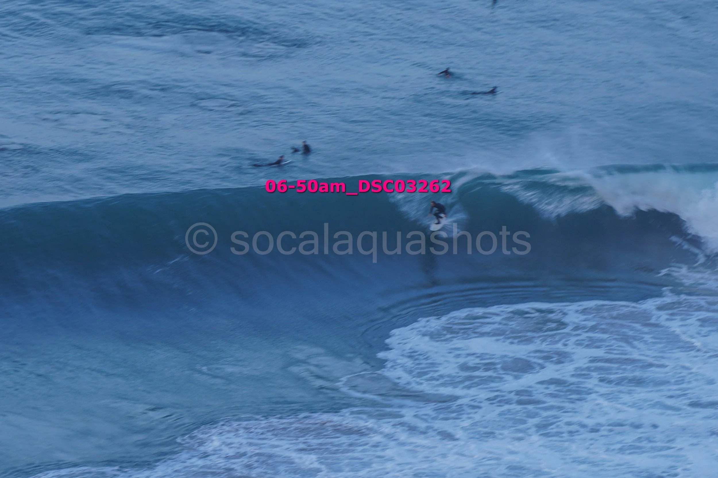 A person surfing on a large wave in the ocean, with several other surfers visible in the background.