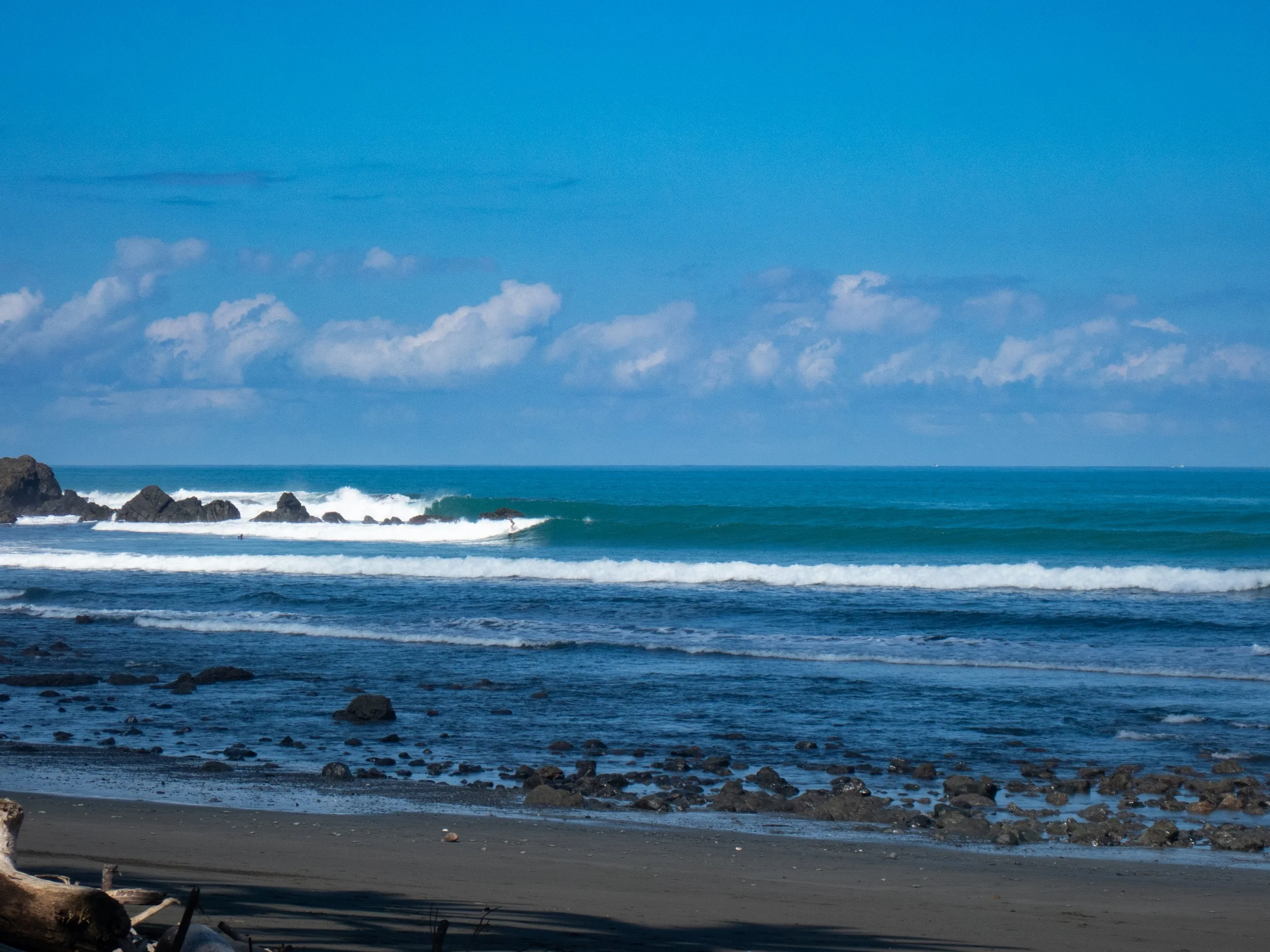 View of the ocean with waves crashing against rocks near the shore, under a blue sky with some scattered clouds.