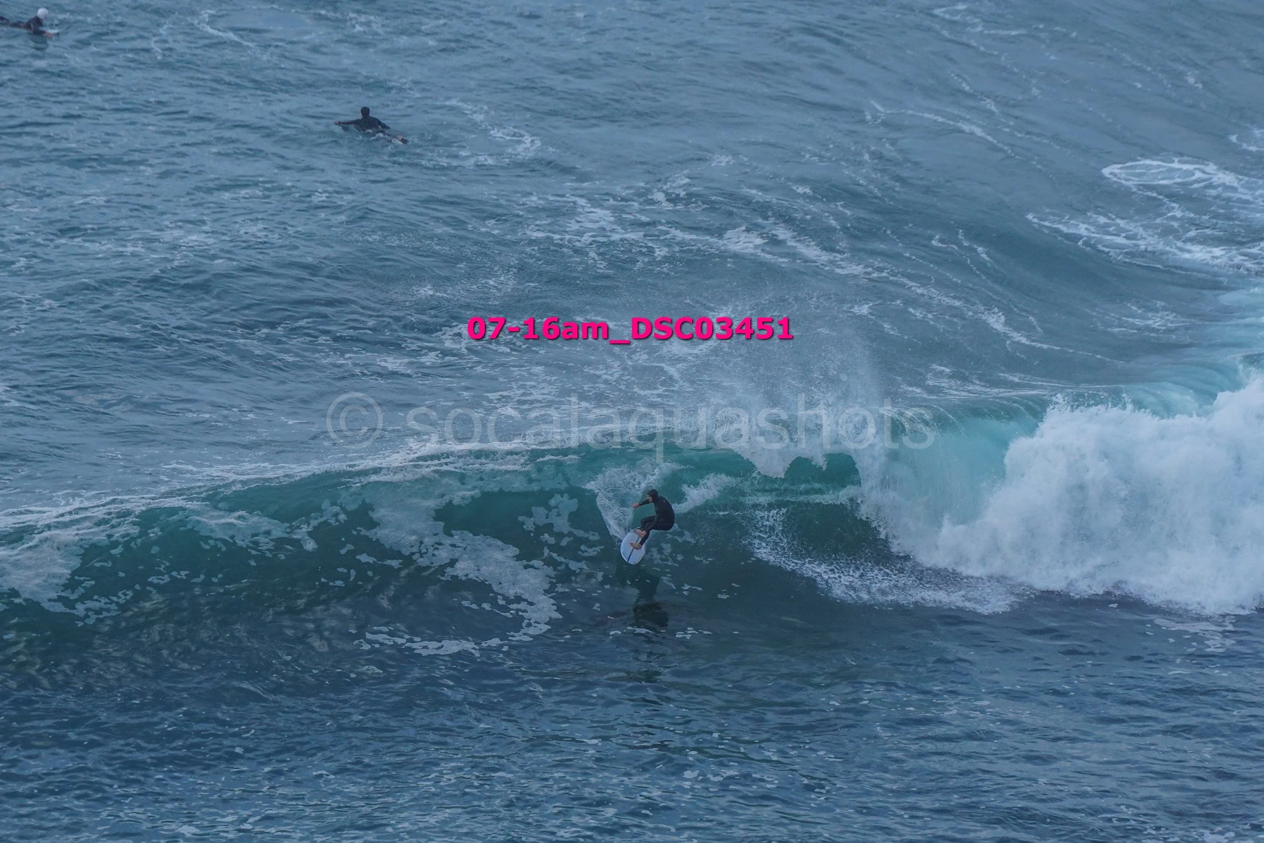 A person surfing on a wave in the ocean during daylight, with another person swimming in the water nearby.